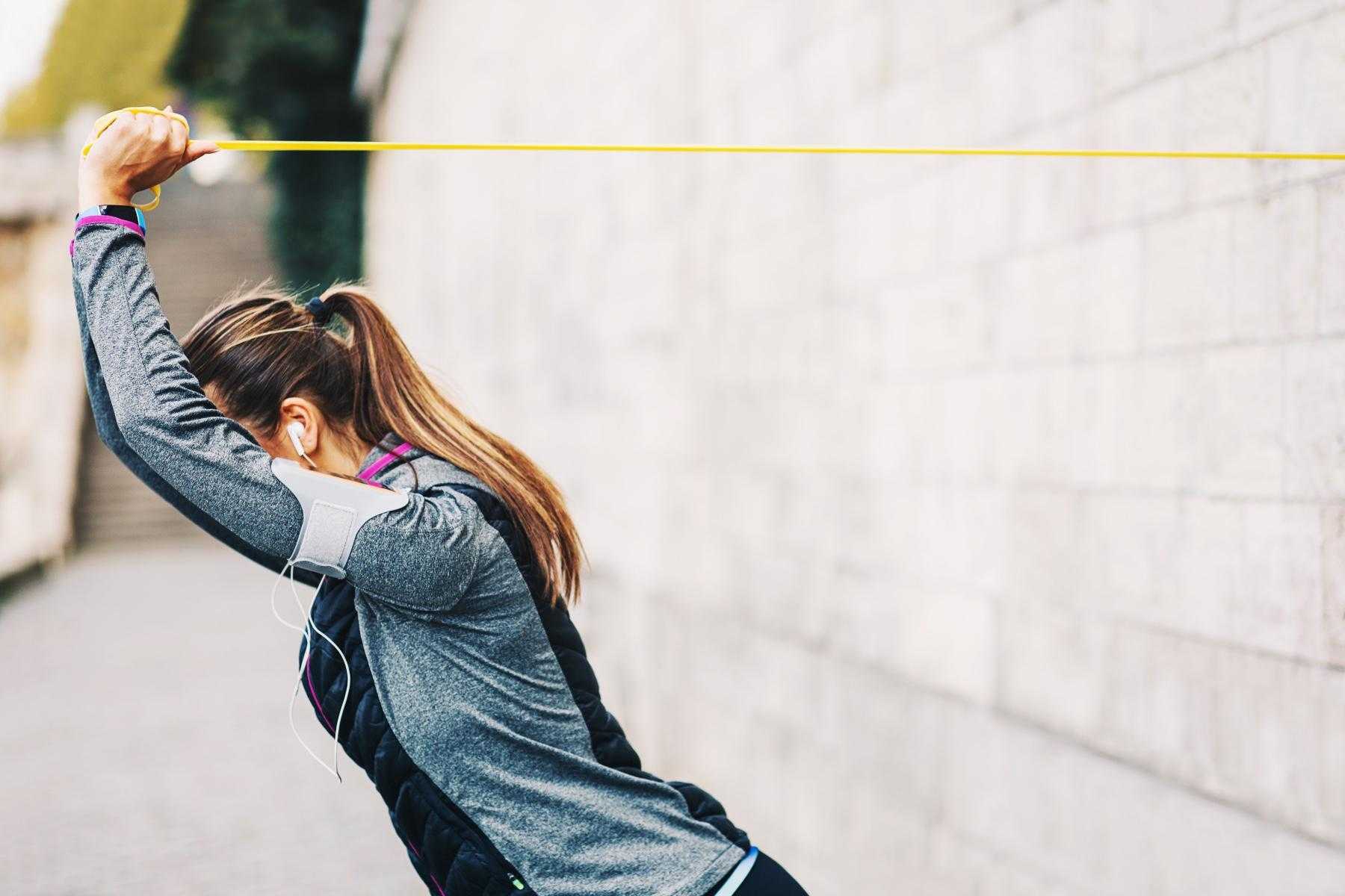 woman in workout clothes outside pulling a yellow resistance band over her head from the wall on the right side