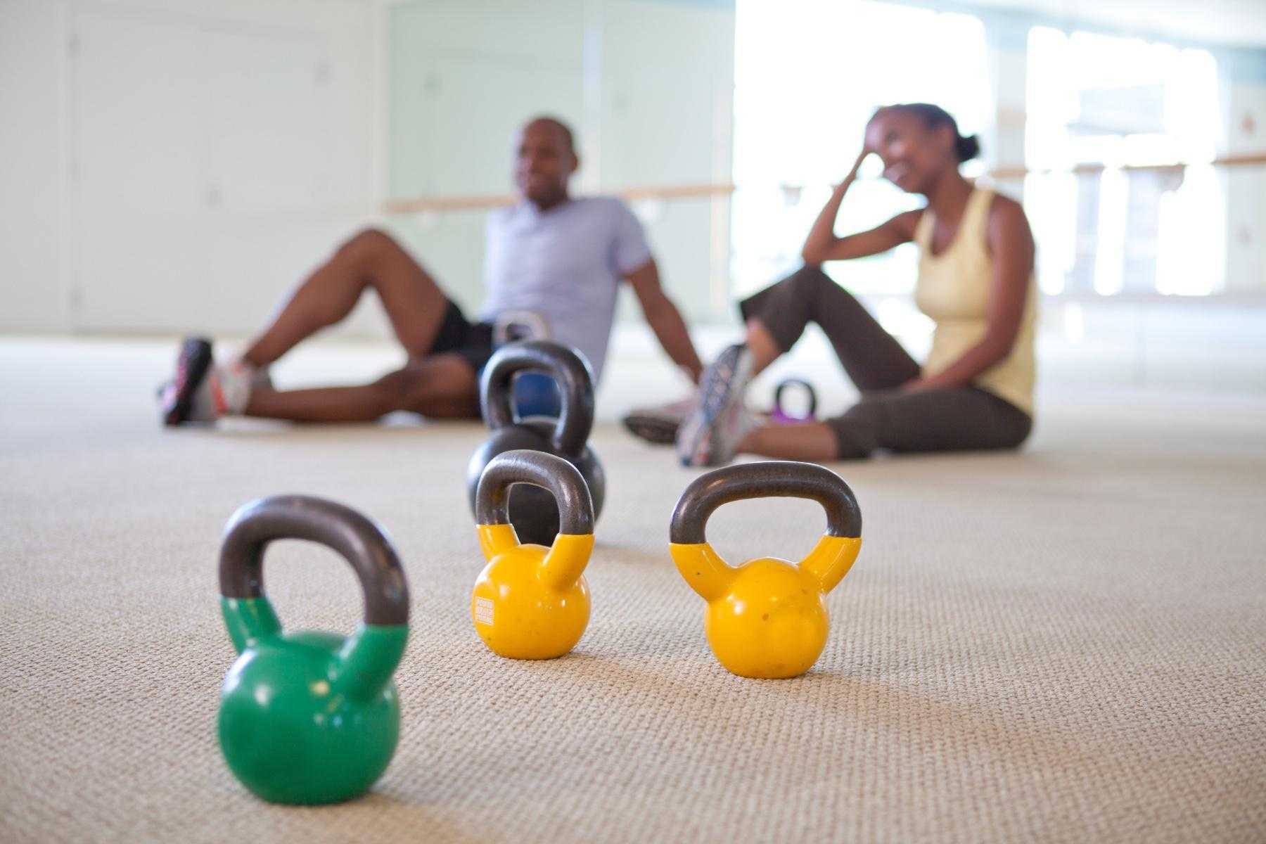 focus on bright yellow and green kettlebells sitting on workout studio floor with a blurry man and woman sitting on the ground in the background smiling
