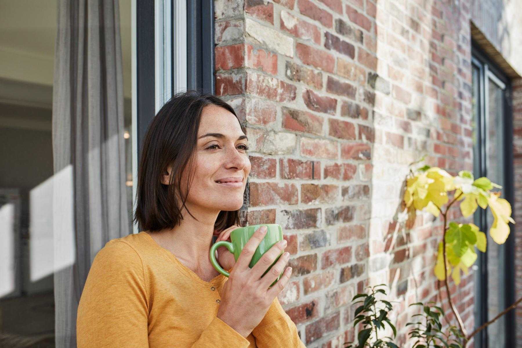 Dark-haired young woman standing outside with back against brick building smiling and holding a cup of tea.