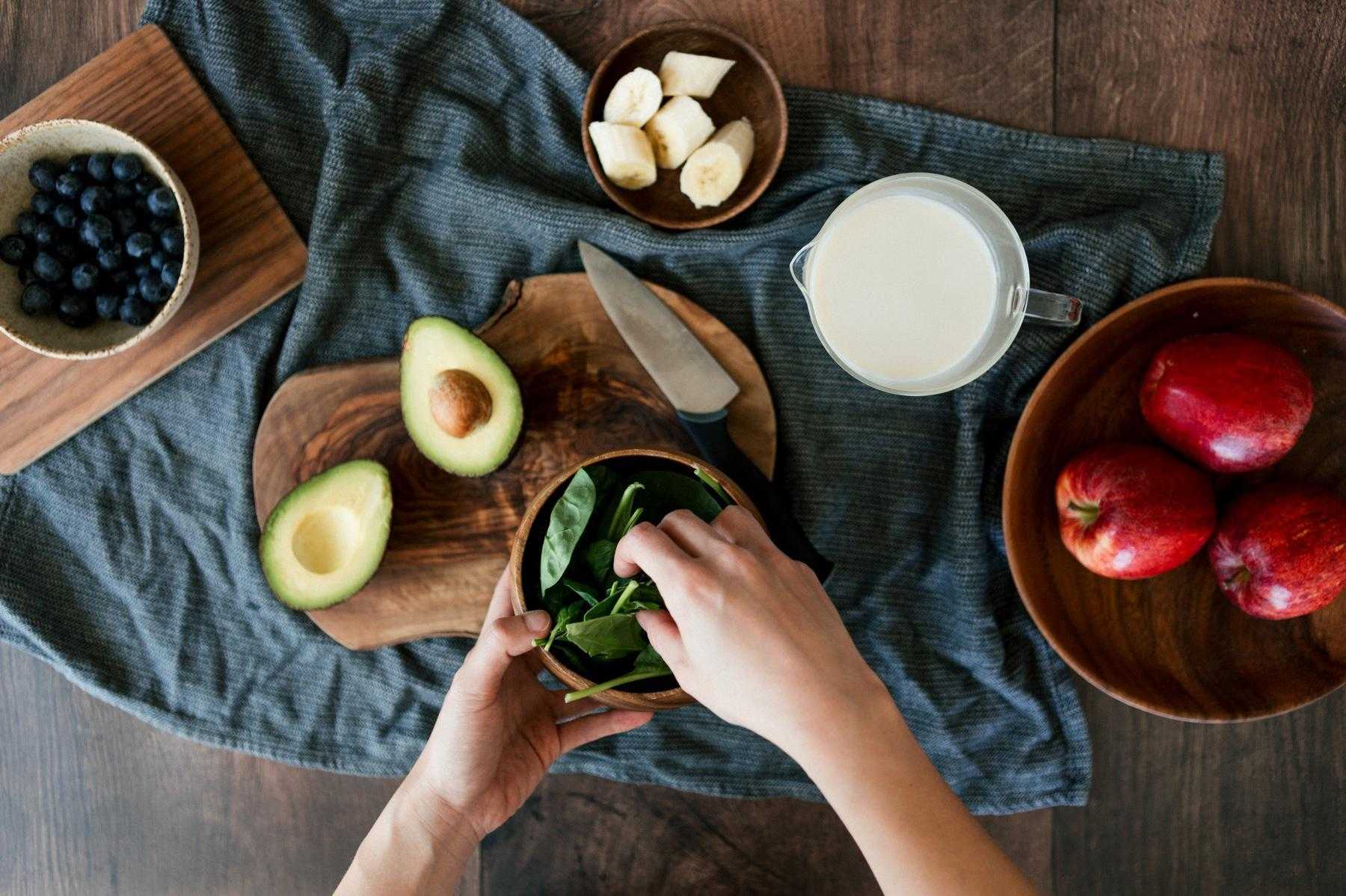 Overhead view of hands reaching over a cutting board with an avocado on it, into bowl of spinach on a table with a bowl of blueberries, apples, and bananas.