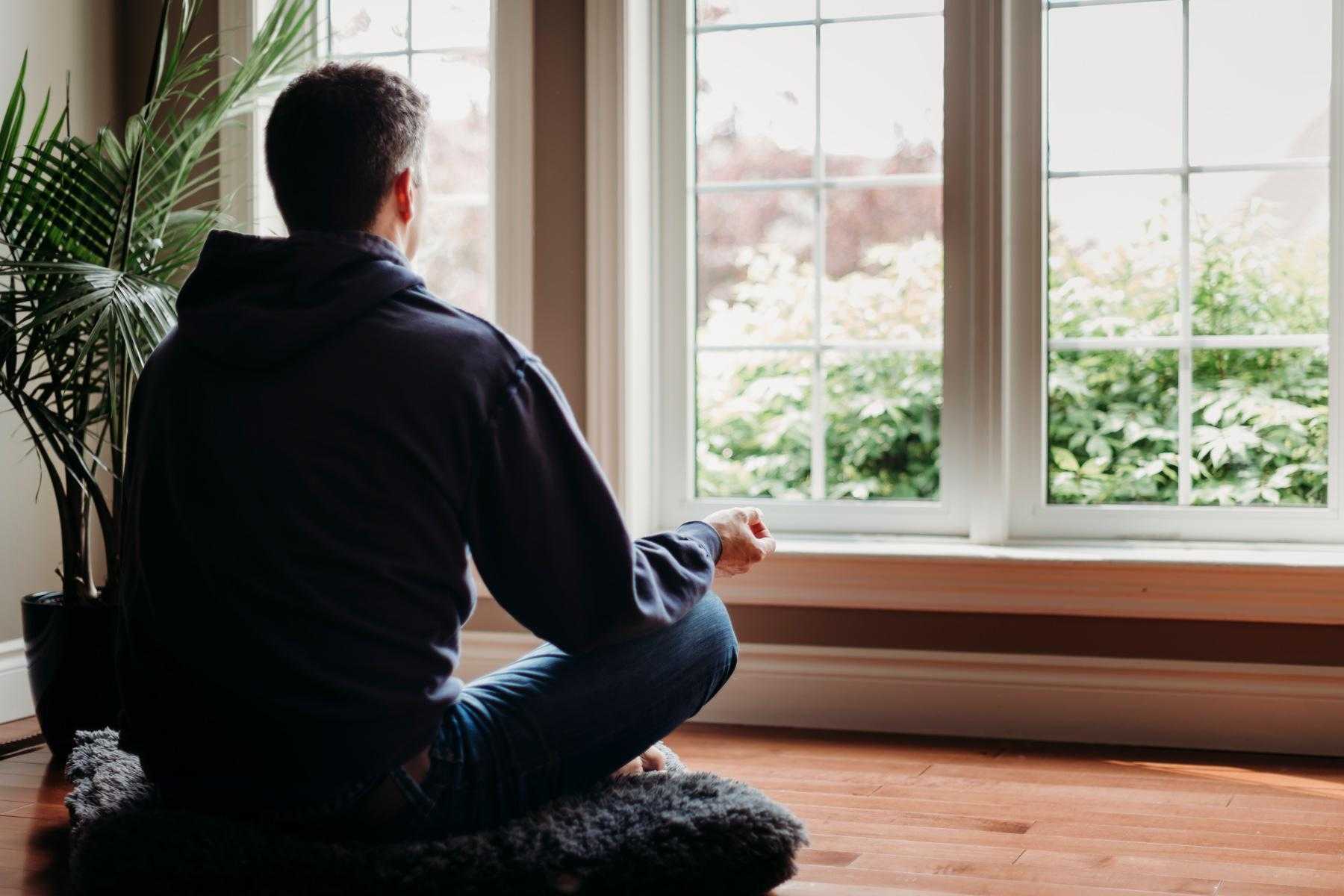 A young man meditating in his living room as he looks out the window.