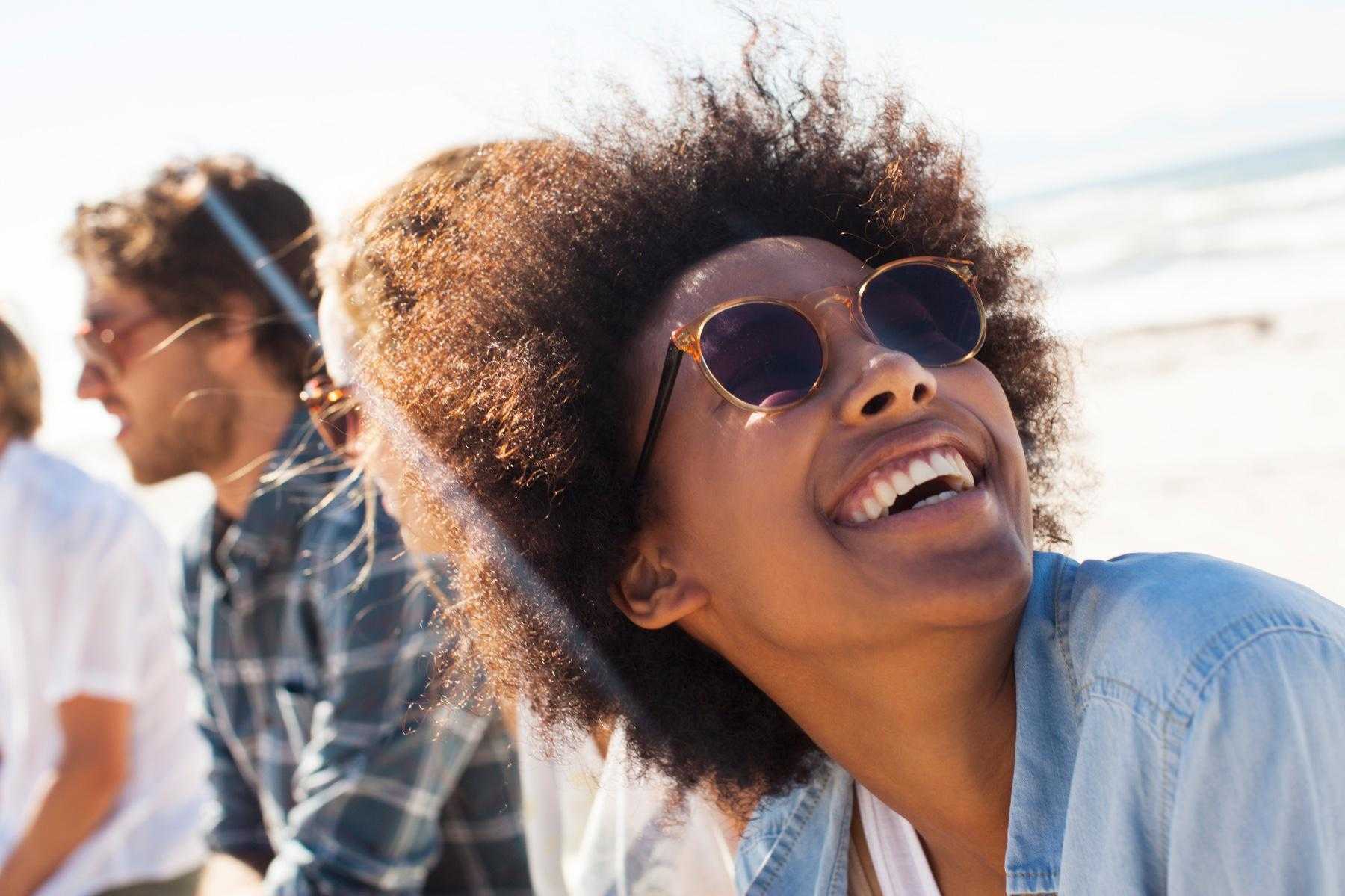 Woman outside with sunglasses on smiling while looking up at the sky with friends in the background.