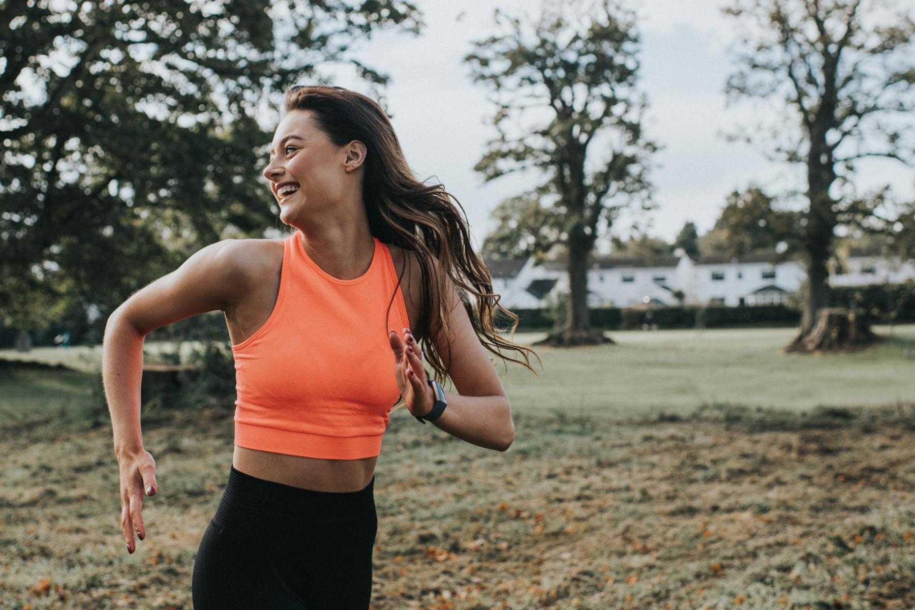 young woman in orange workout shirt running outside while looking behind her and laughing