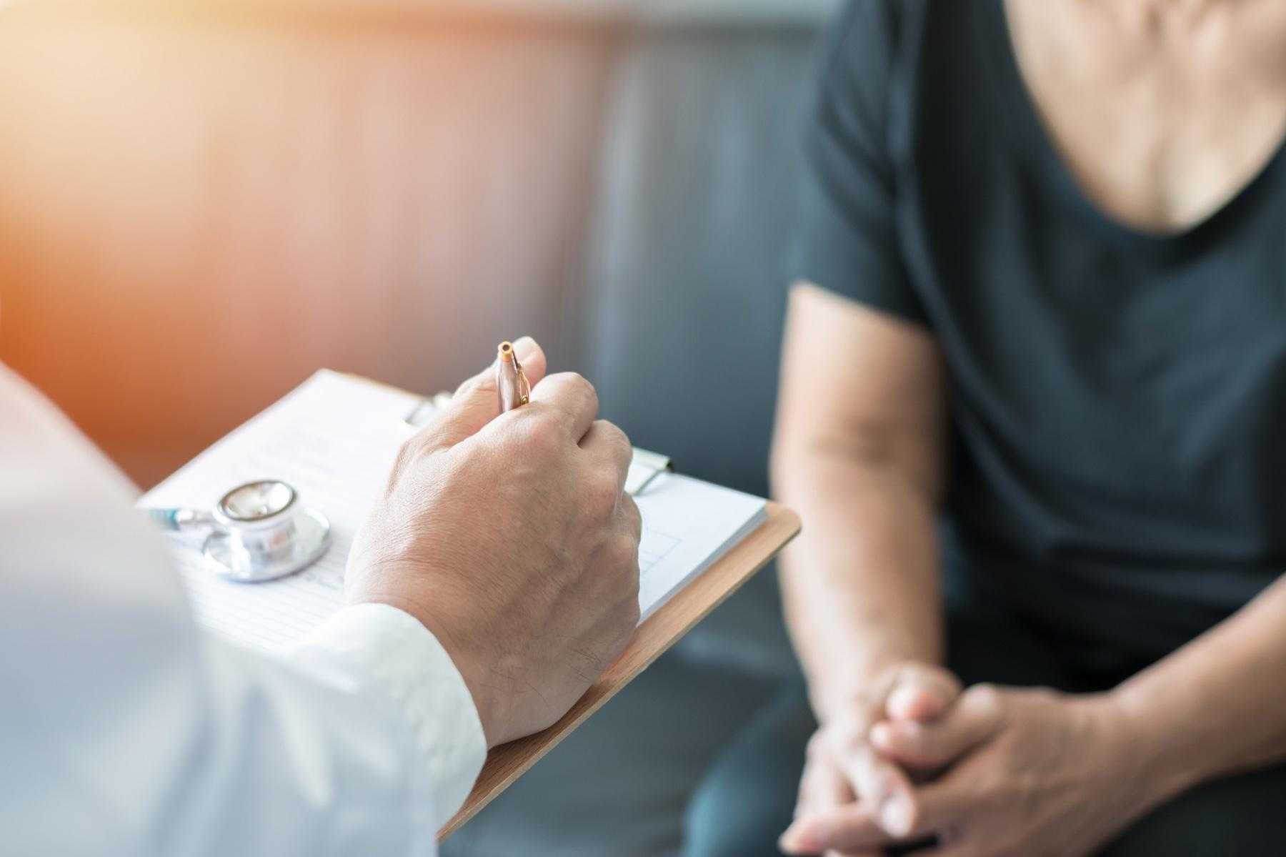Closeup of doctor writing notes on clipboard as patient sits in the background.