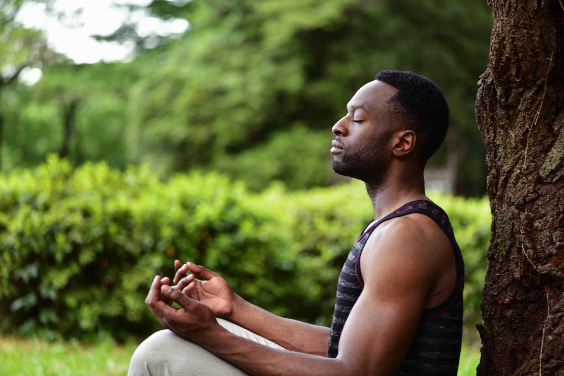 A young man meditating outside in a park next to a tree.