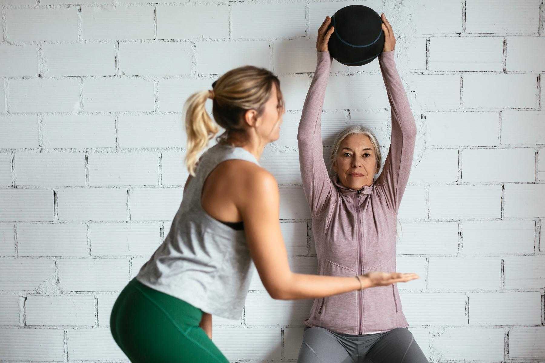one woman spotting another woman as she does a wall sit against a white brick wall while holding a medicine ball above her head