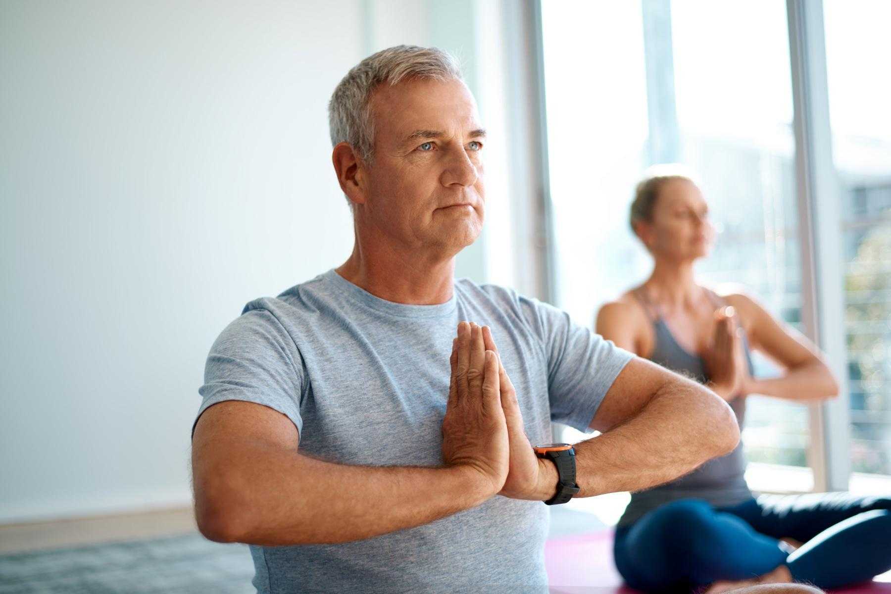 Close-up of an older man with hands at heart center, older woman blurry in the background with hands also at heart center.