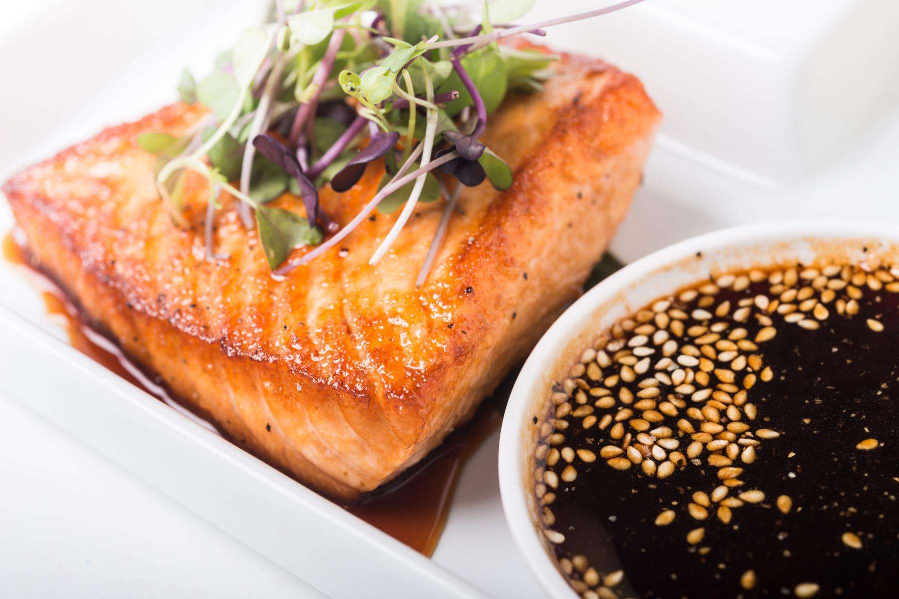 Cooked salmon on a plate next to a dish of teriyaki sauce.