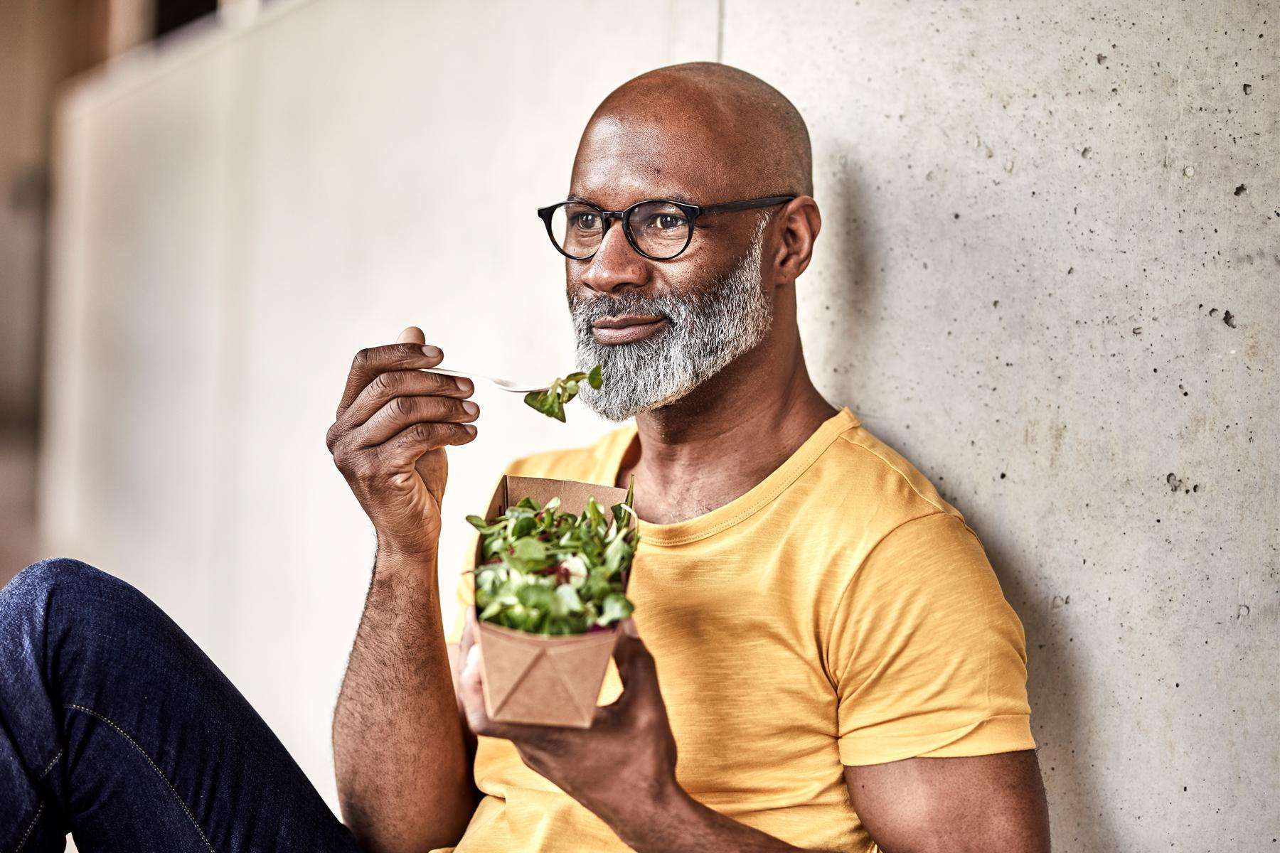 Image of a middle-aged man sitting up against a wall while eating a salad.