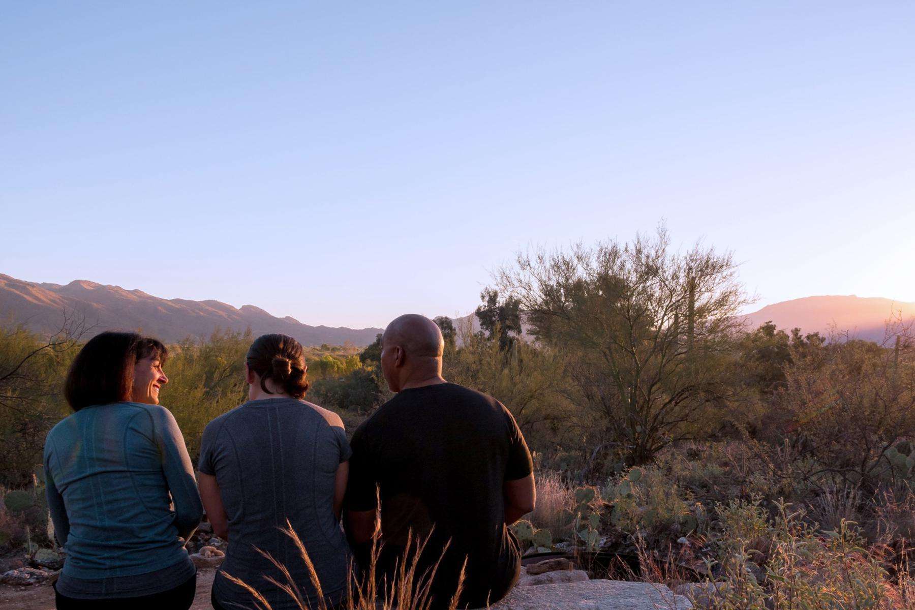 In the desert, there are three friends sitting on a large rock laughing as the sky turns to dusk.
