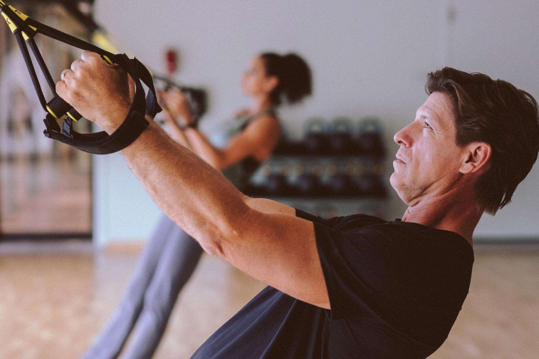 Man and woman doing rows on TRX straps in gym.