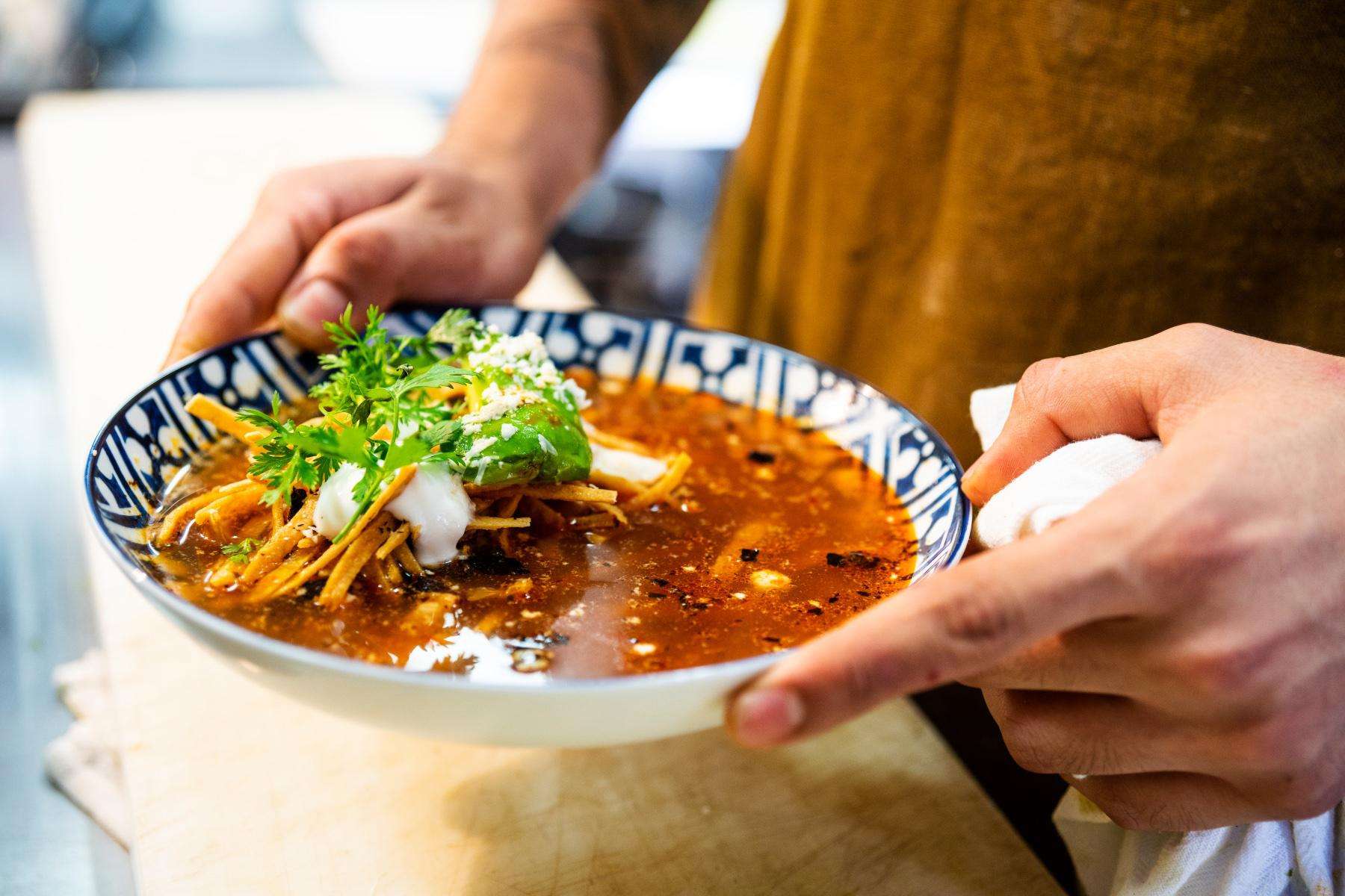 Hands holding a bowl of tortilla soup.