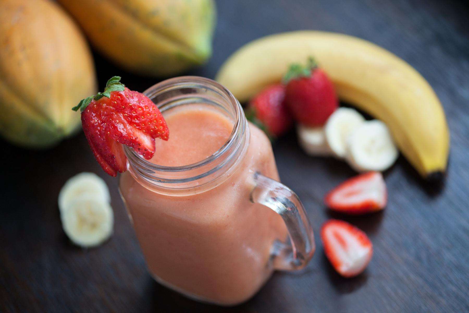 A countertop with strawberry and banana pieces and a mason jar filled with a strawberry banana smoothie.