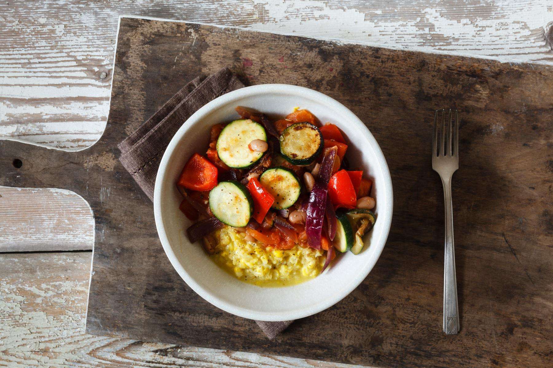 Overhead view of a bowl filled with grilled polenta and marinated vegetables on wooden cutting board.