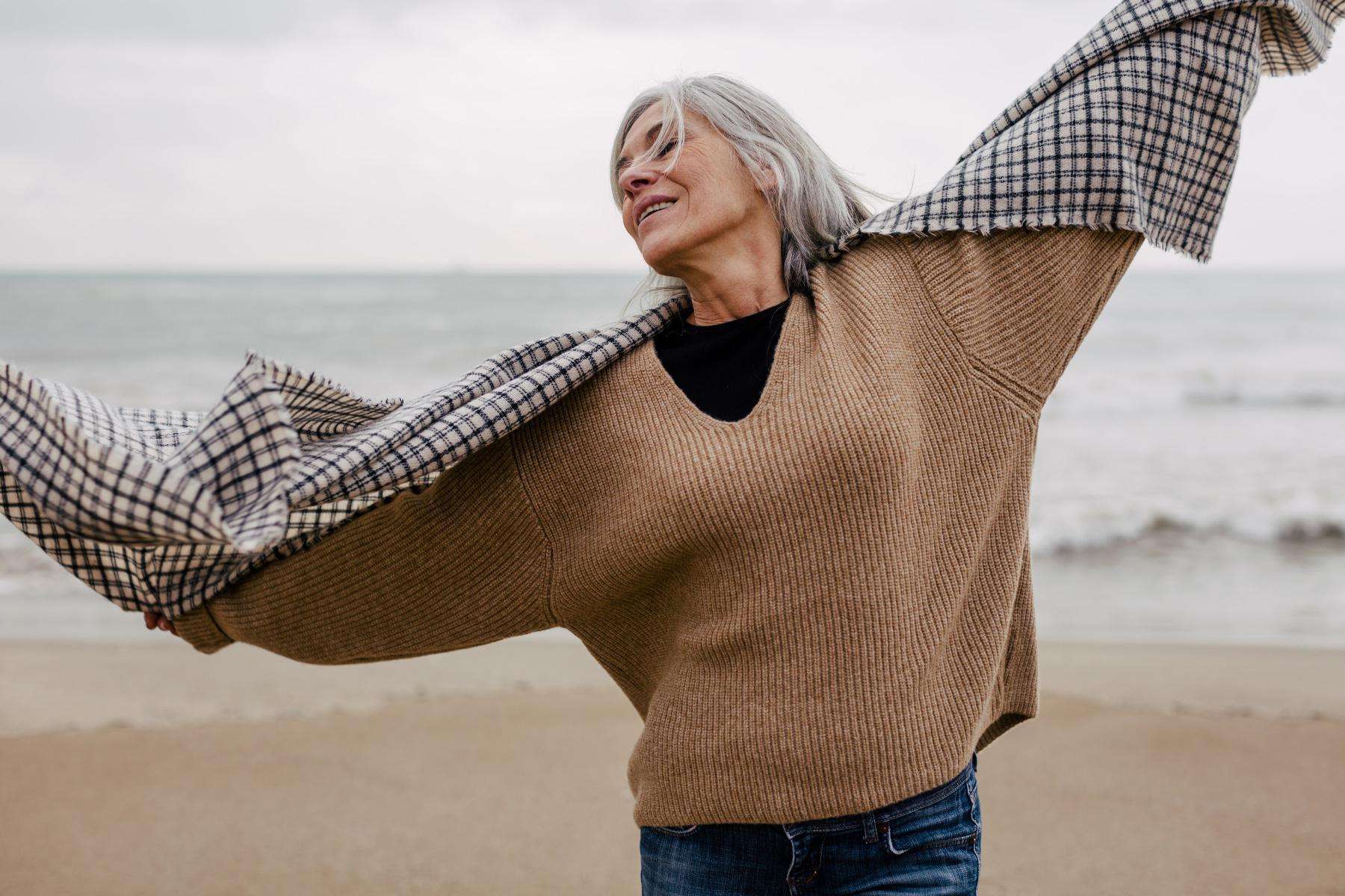 Older woman by ocean with scarf wrapped around her arms as she raises them up with her head back and eyes closed.