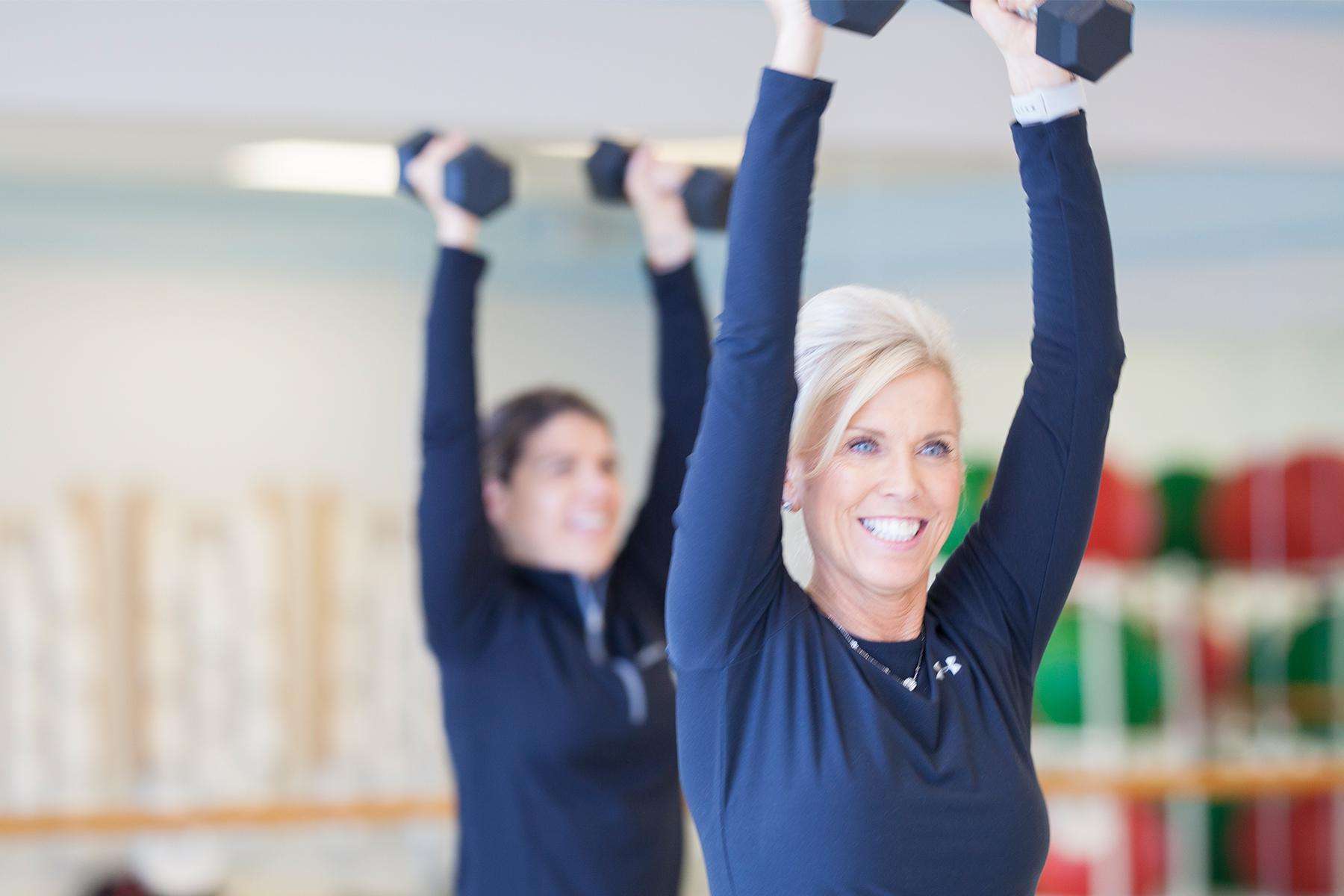 two women wearing all black in fitness room holding dumbbells above their head during class