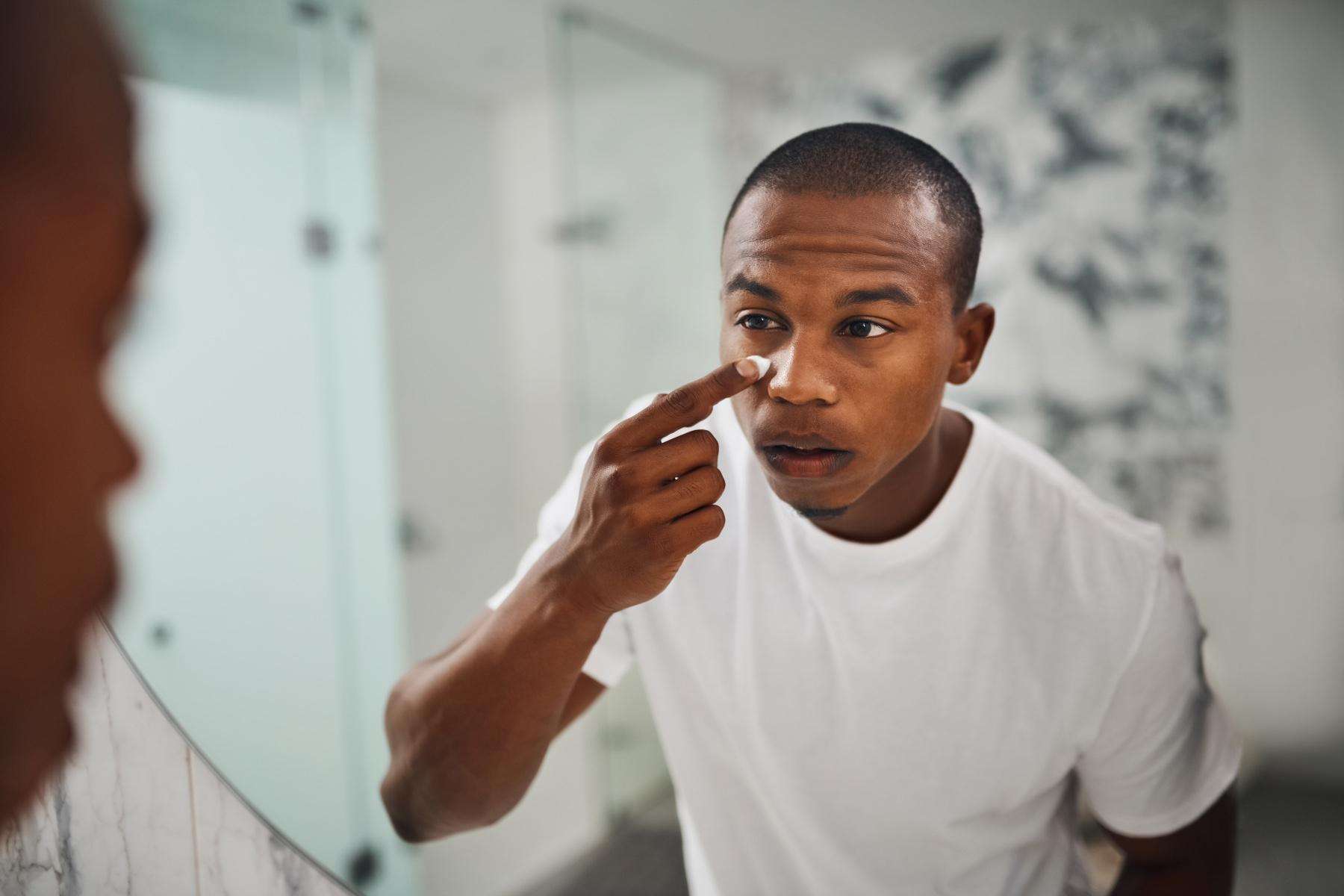 Young man standing in front of his bathroom mirror at home applying lotion to his face.