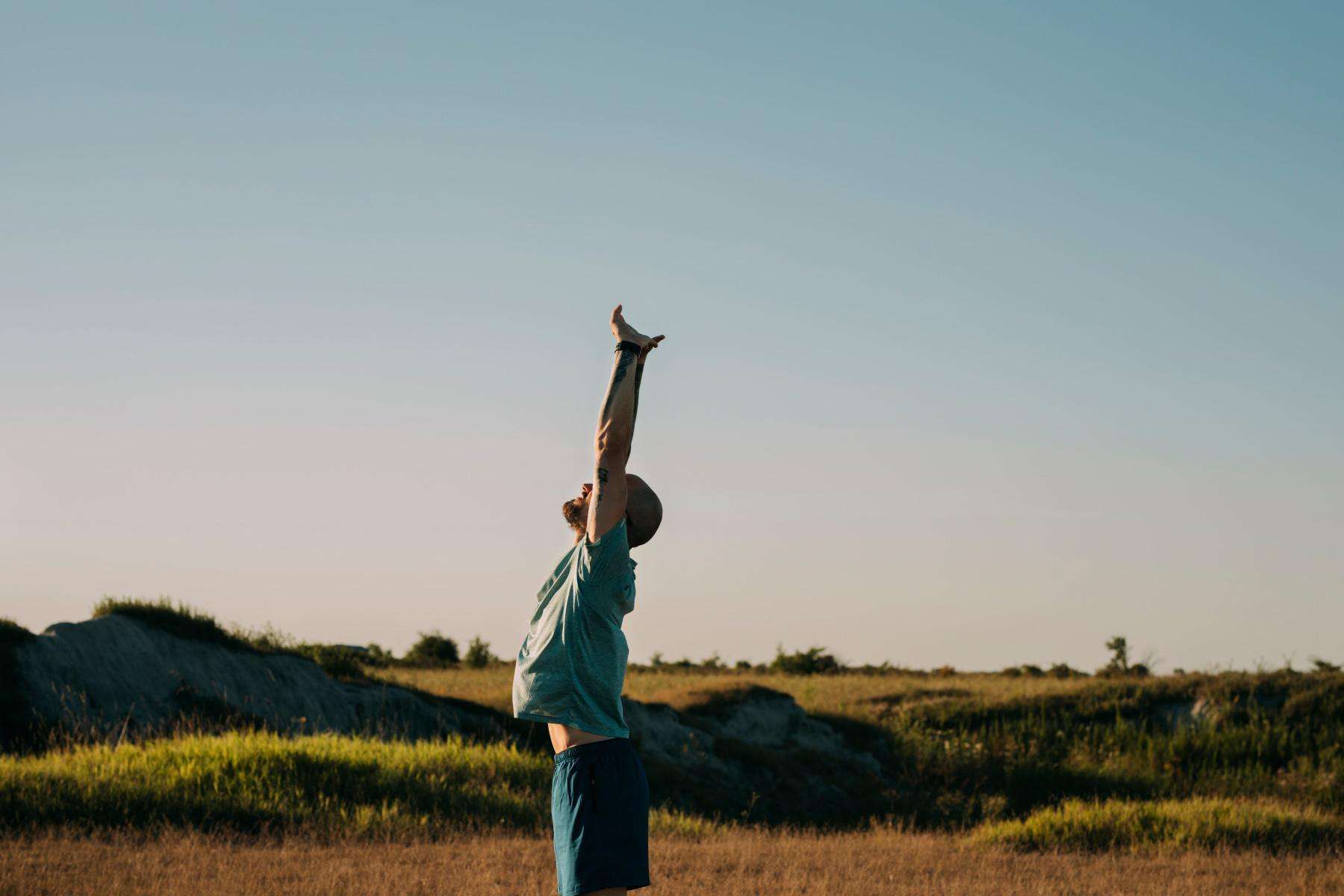 side view of man with arms raised above head stretching outside with a bright blue sky and green grass behind him