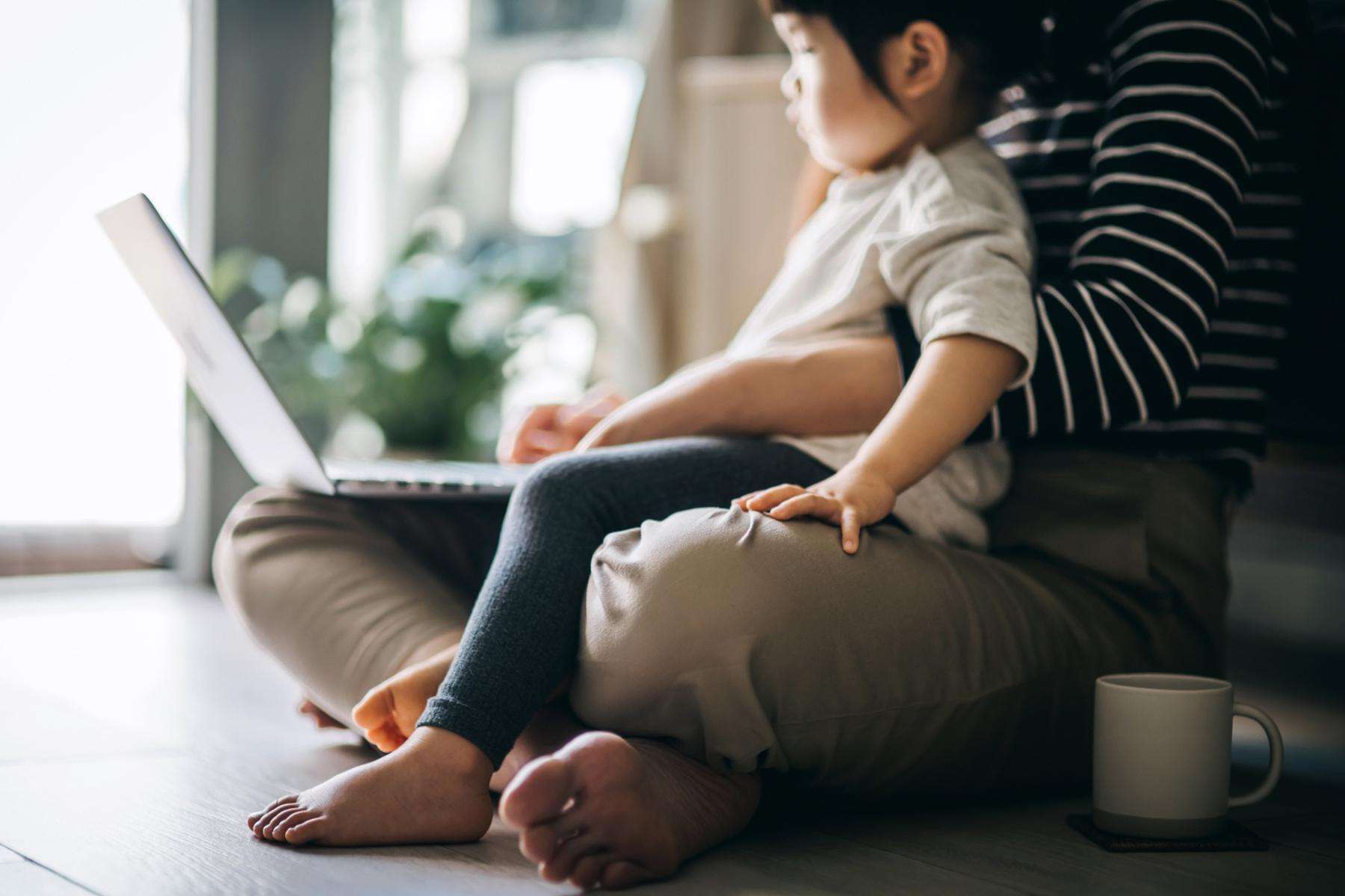 Woman sitting on ground working on laptop with toddler sitting in lap.