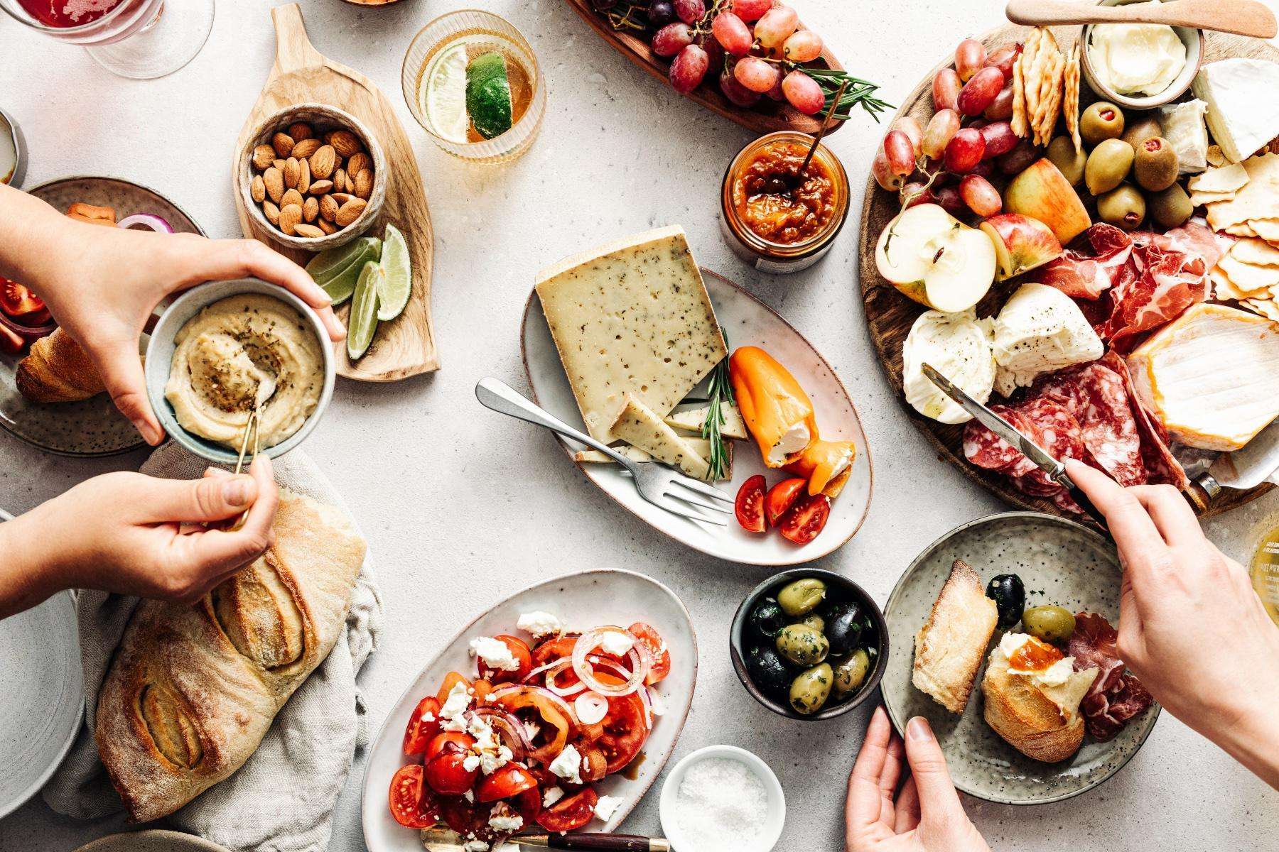 overhead table view of hands serving from plates of mediterranean food including cheese and olives