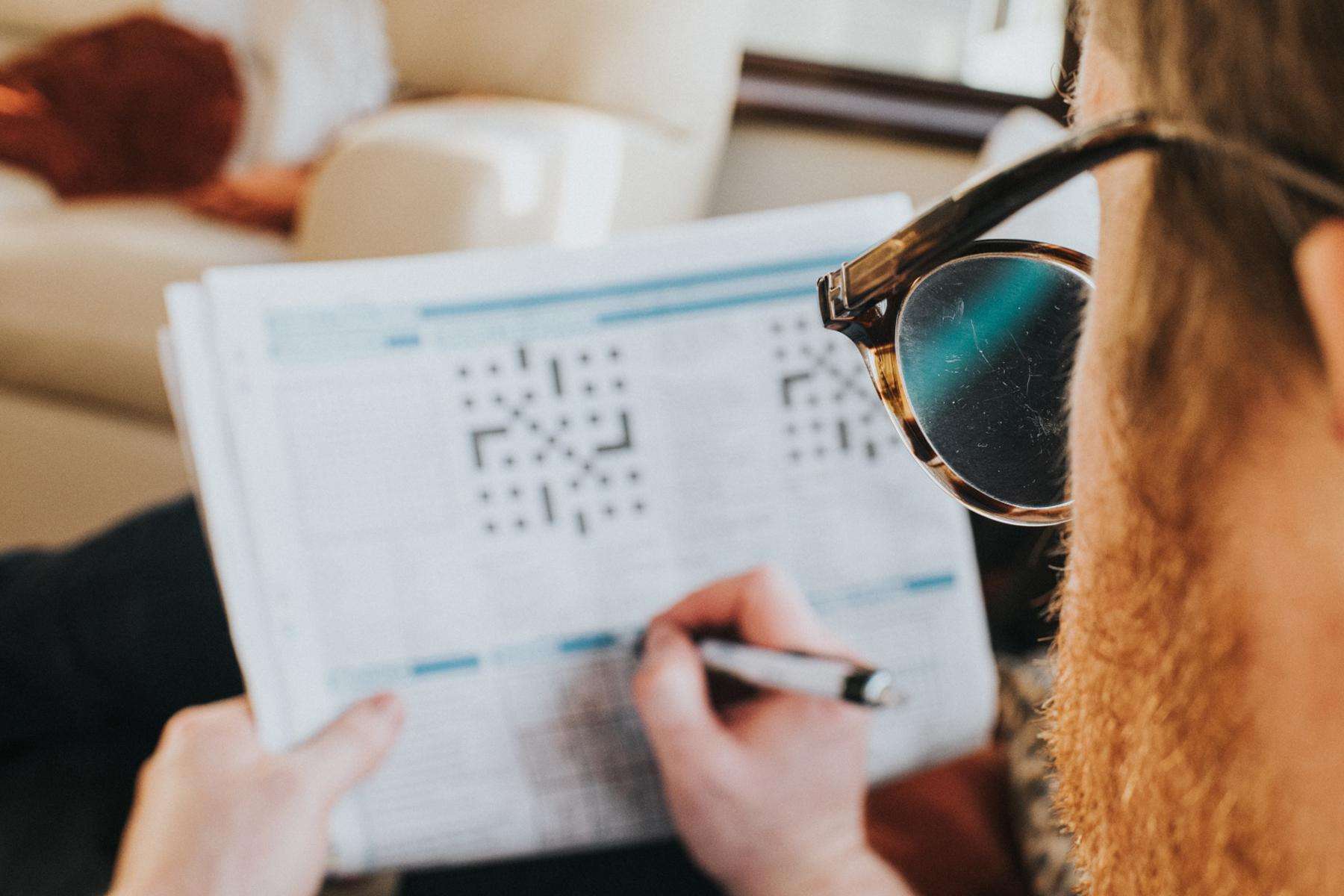Looking over the shoulder of a man with glasses on doing a crossword puzzle in the newspaper while sitting on a couch.