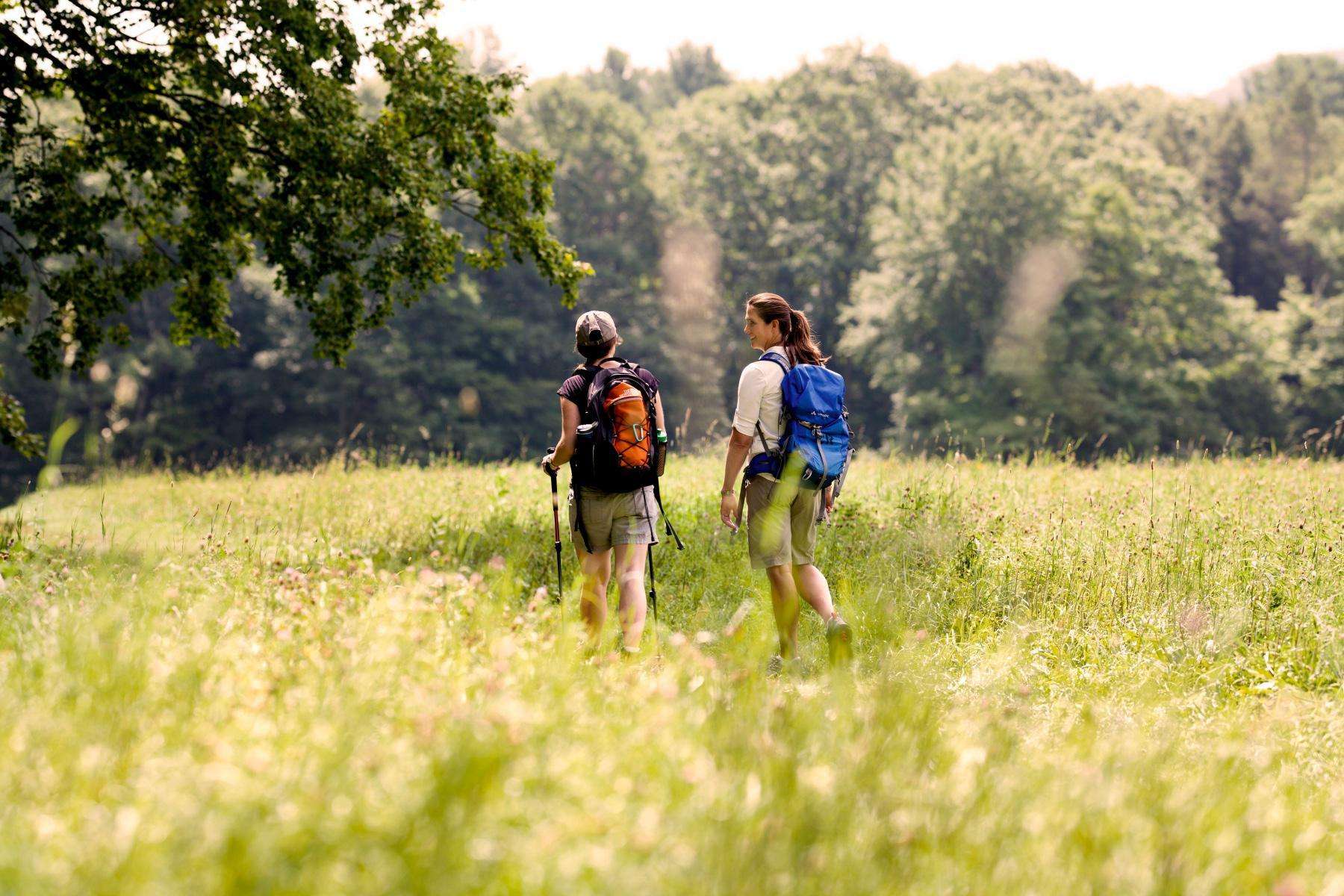 Two women hiking through green field with backpacks on.