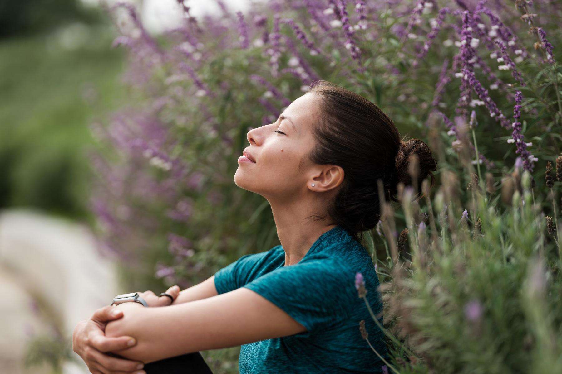 Woman sitting against bush with purple flowers with eyes closed smiling.