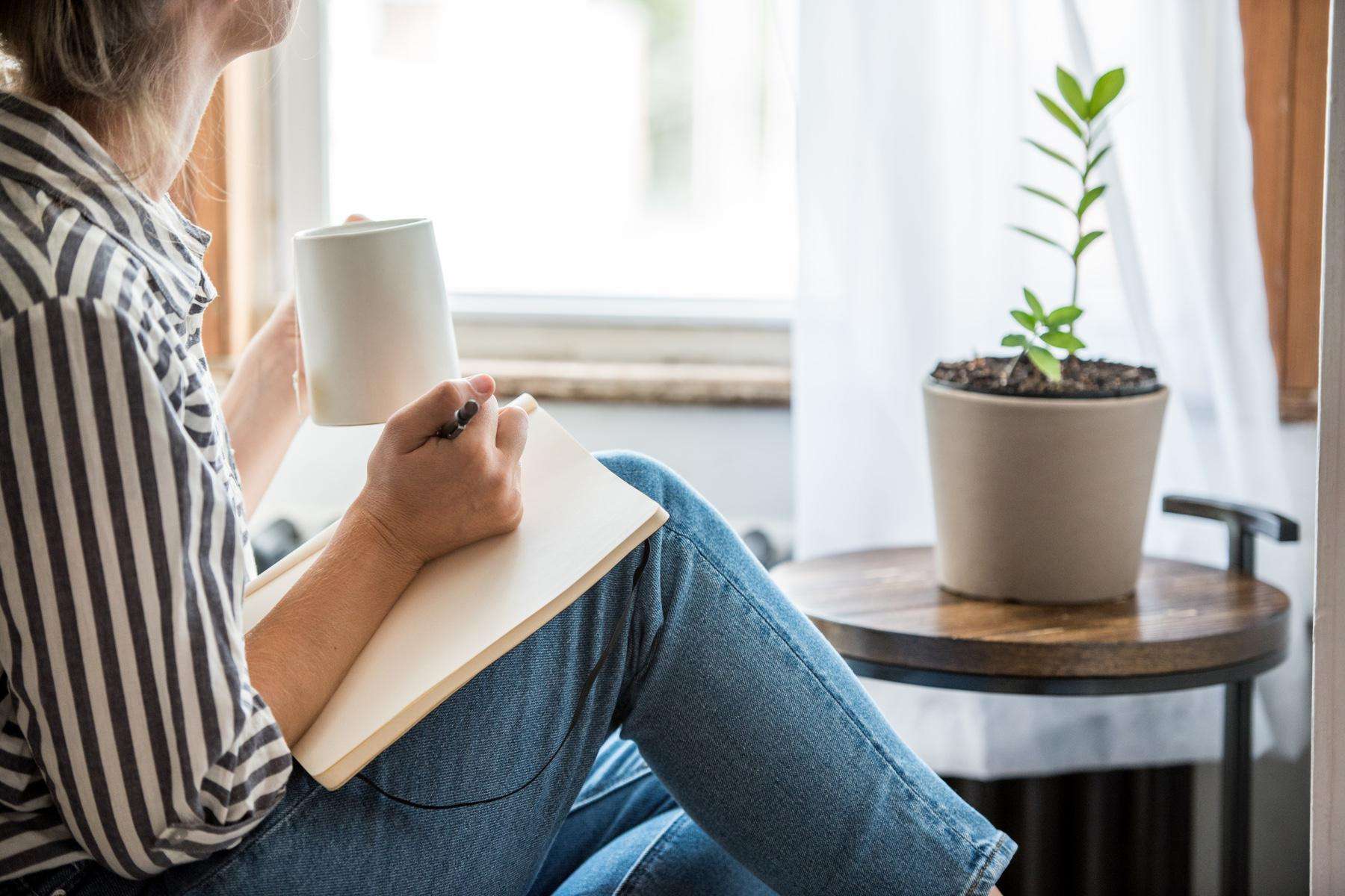 Woman writing in her journal as she sits in her living room with a small potted plant in front of her.