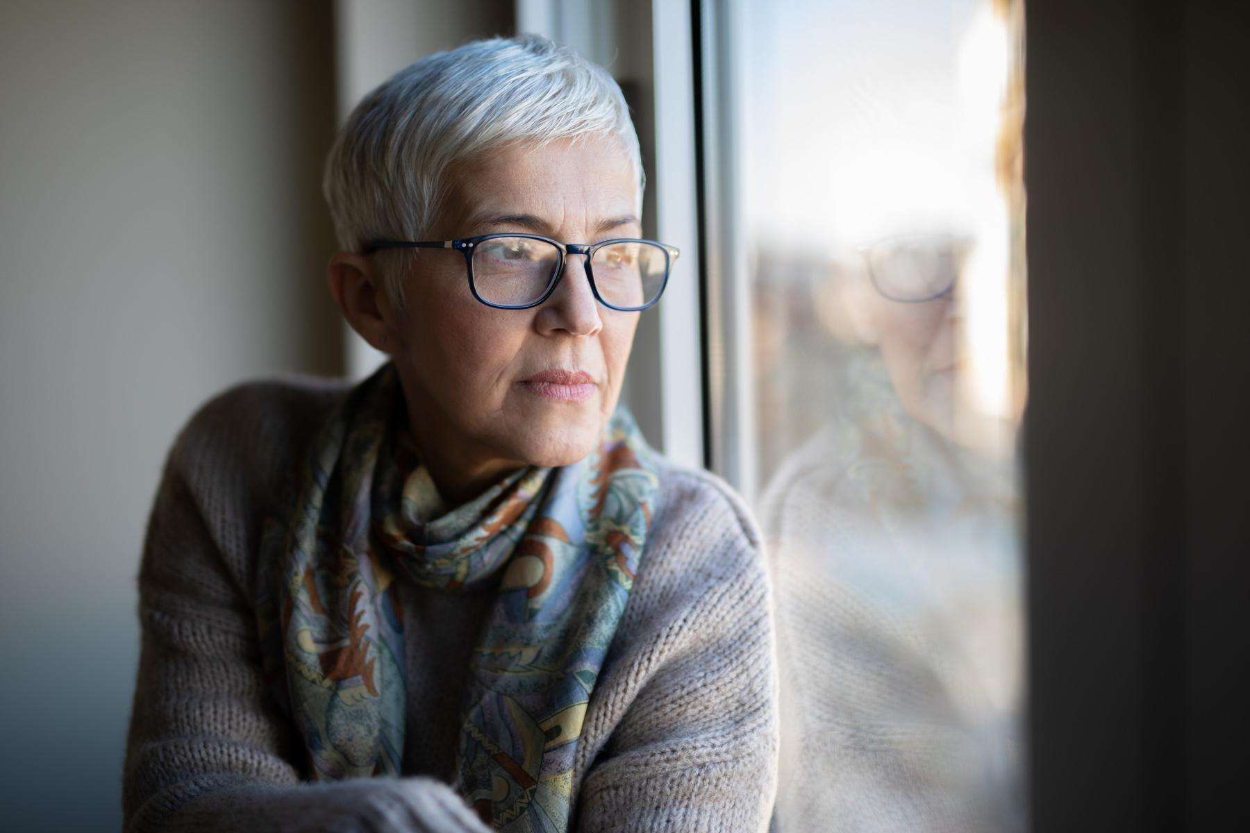 Older woman with glasses sitting up against window looking outside.