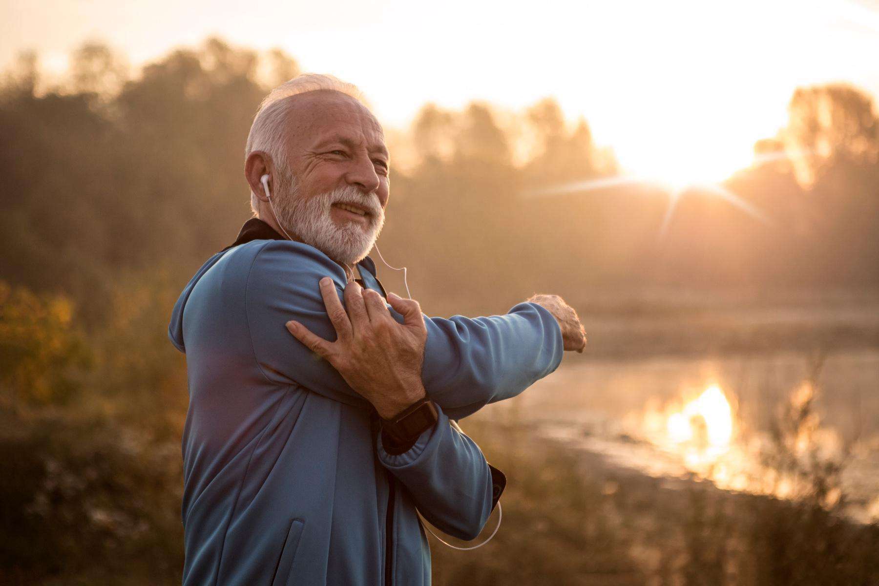 Older man outside with headphones on stretching his arm across his chest as the sun rises.