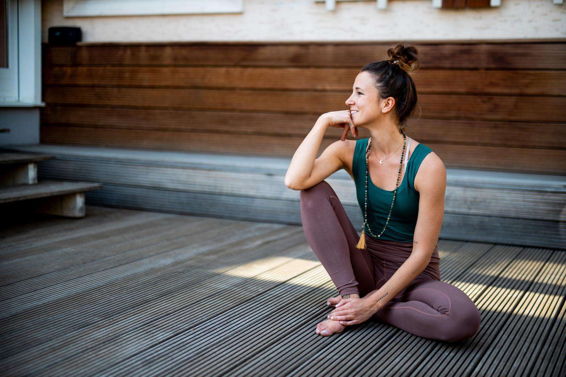 Woman sitting outside on floor of deck after yoga practice smiling and looking off to the side.