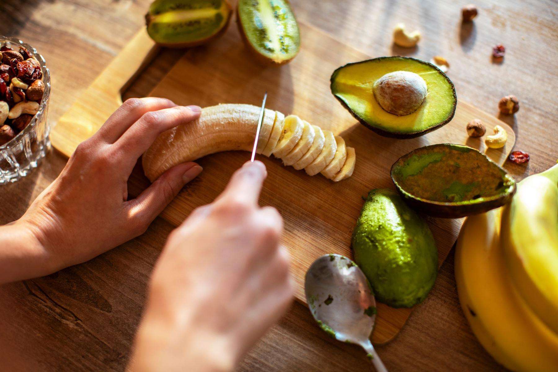 Close-up of hands chopping a banana with a knife on a wooden cutting board.