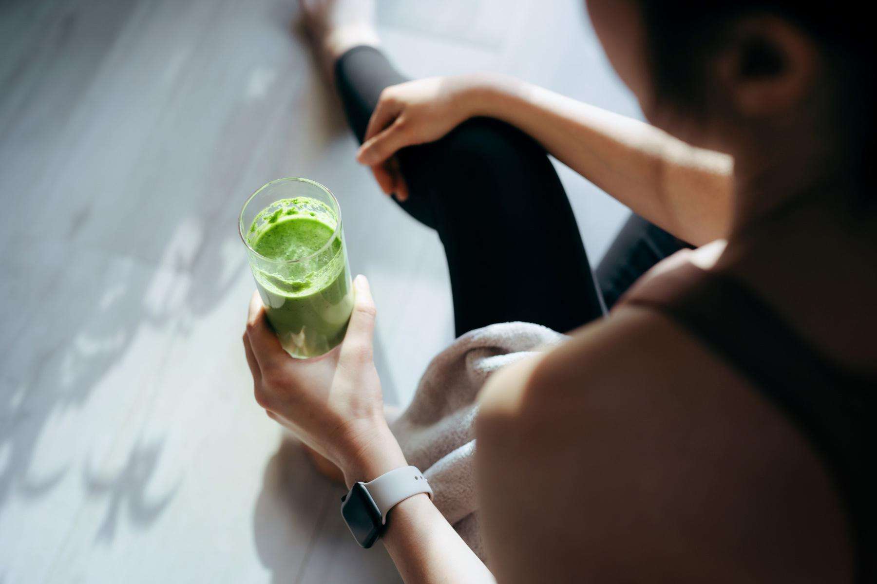 Woman sitting on the ground after working out drinking a green smoothie.
