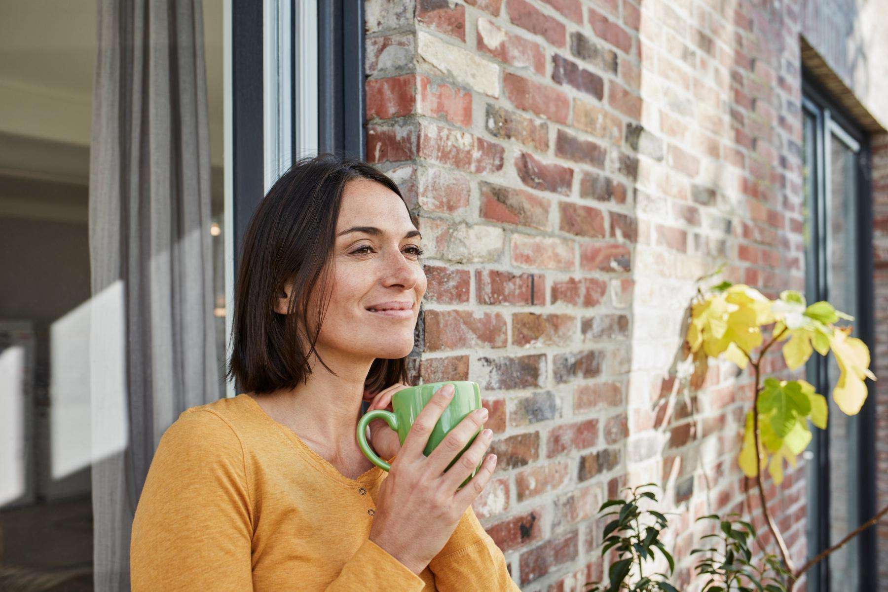 Dark-haired young woman standing outside with back against brick building smiling and holding a cup of tea.