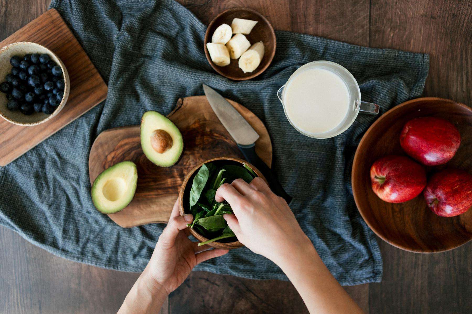Overhead view of hands reaching over a cutting board with an avocado on it, into bowl of spinach on a table with a bowl of blueberries, apples, and bananas.