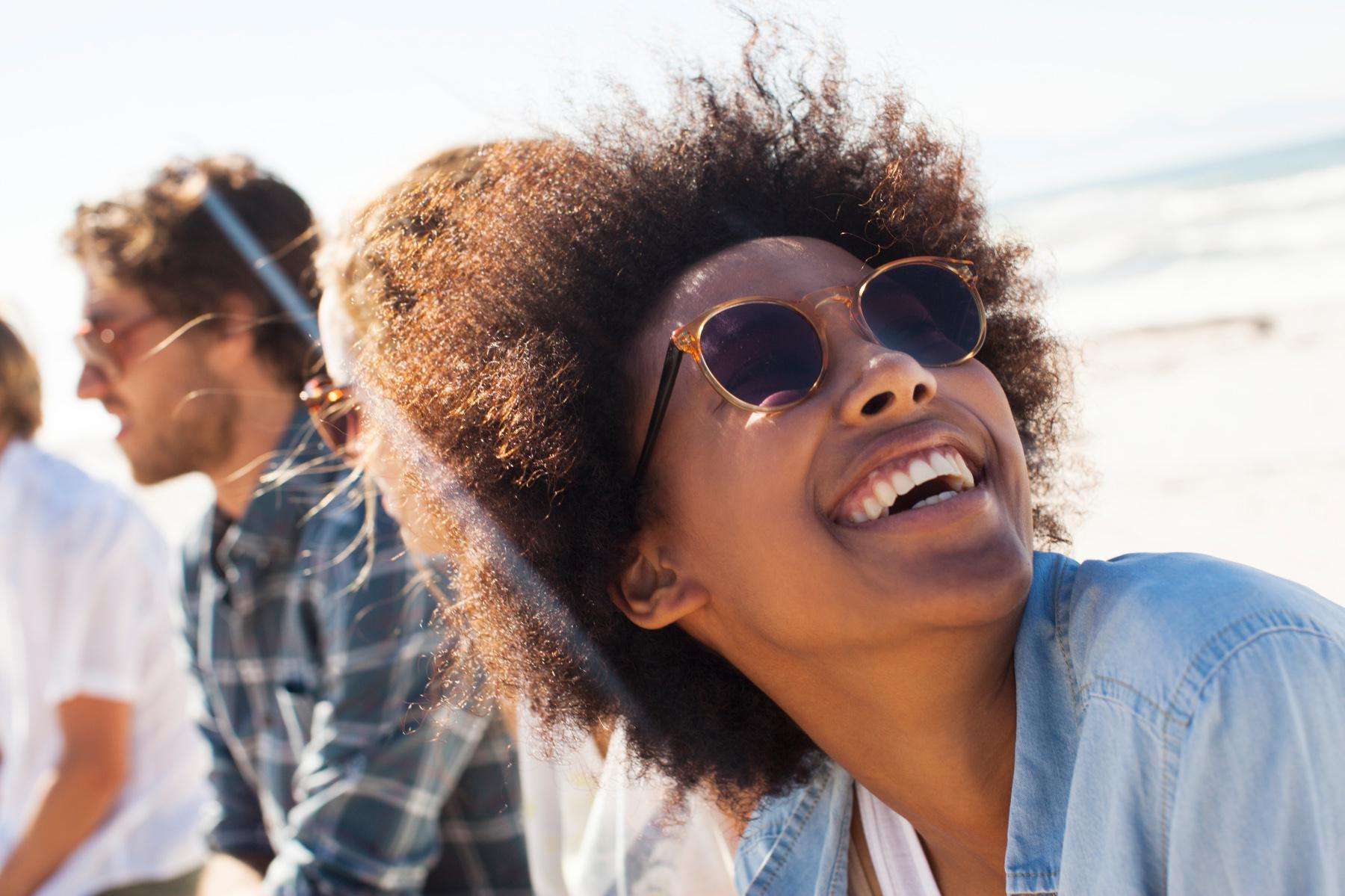 Woman outside with sunglasses on smiling while looking up at the sky with friends in the background.