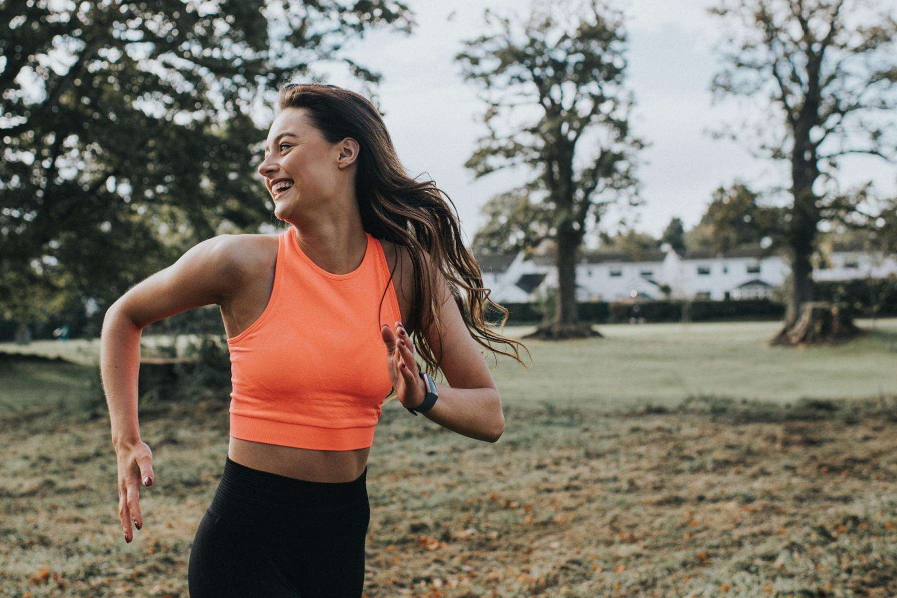 young woman in orange workout shirt running outside while looking behind her and laughing