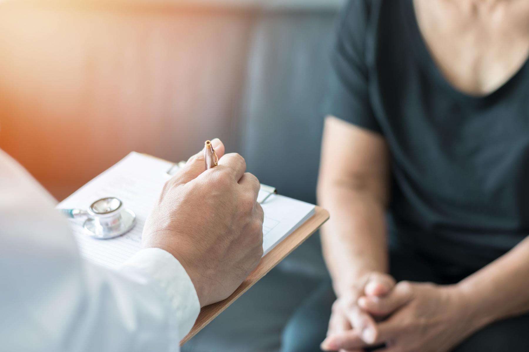 Closeup of doctor writing notes on clipboard as patient sits in the background.