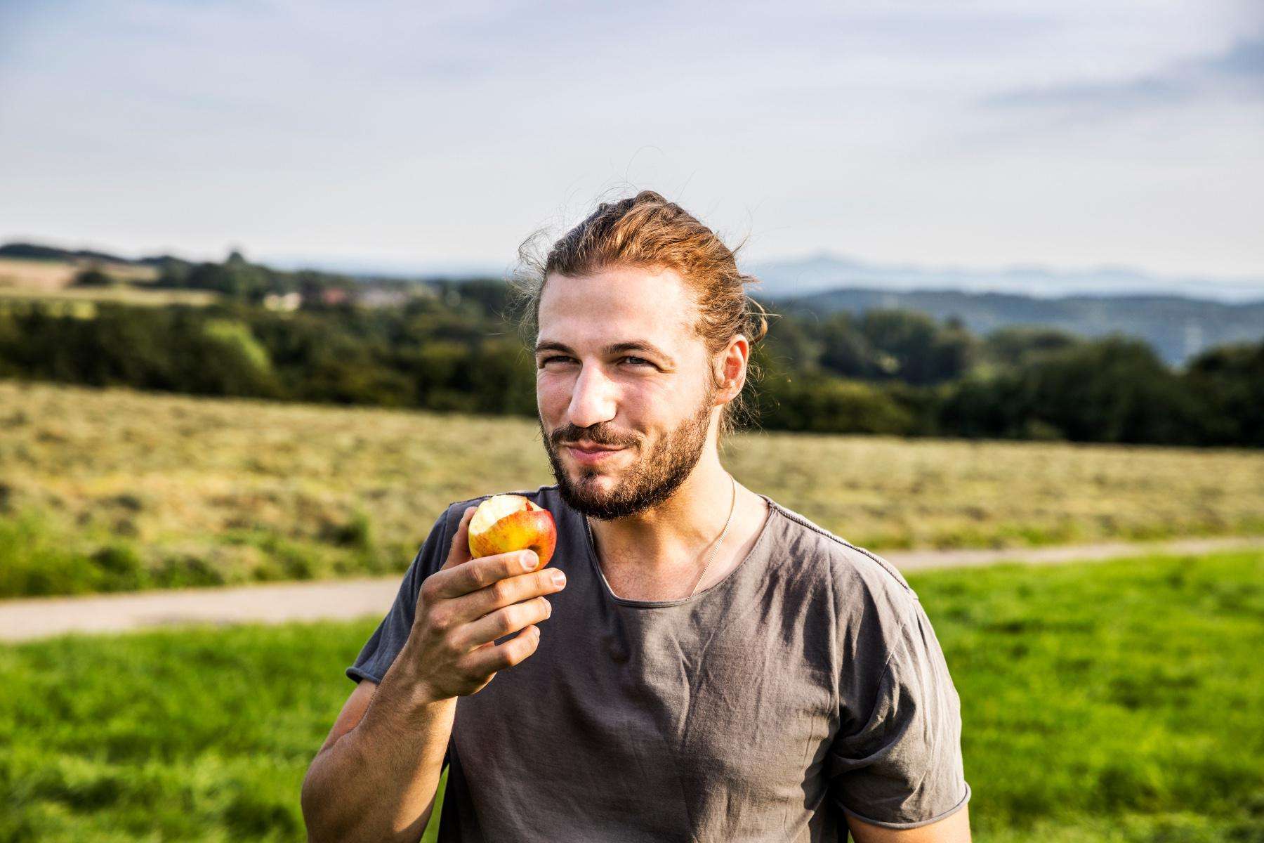 Man eating red apple outside with green grass and mountain backdrop.