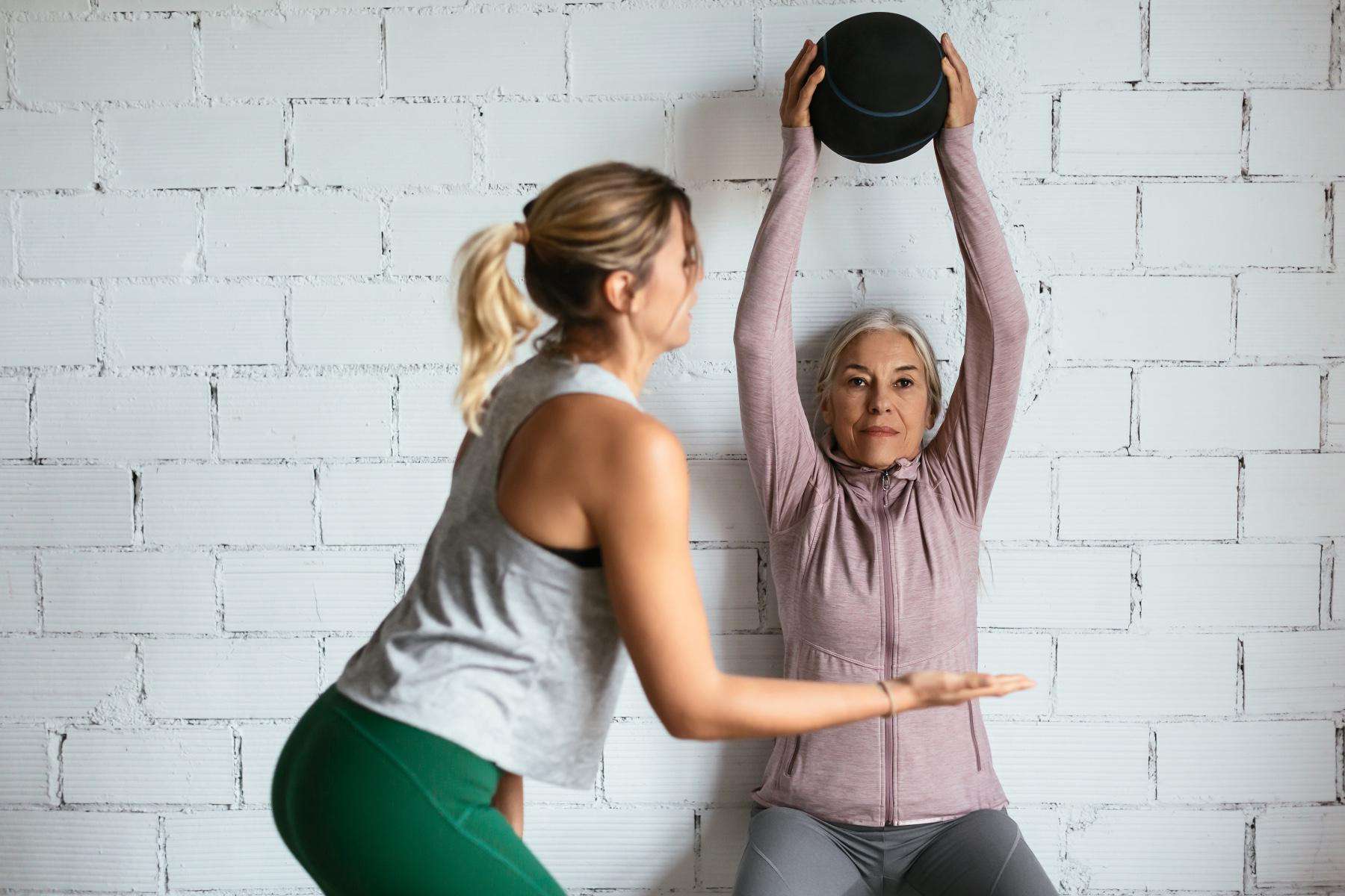 one woman spotting another woman as she does a wall sit against a white brick wall while holding a medicine ball above her head