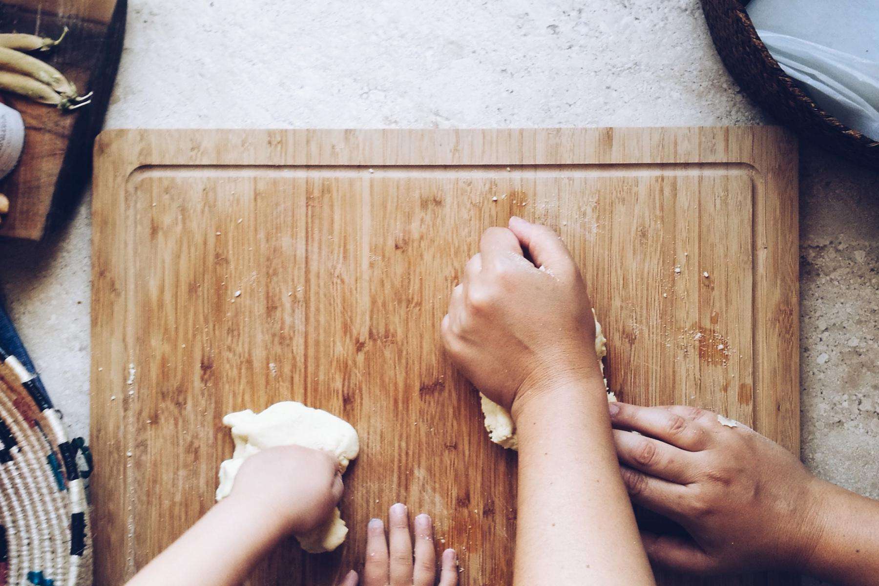 overhead image of woman's hands and child's hands rolling dough on wooden cutting board