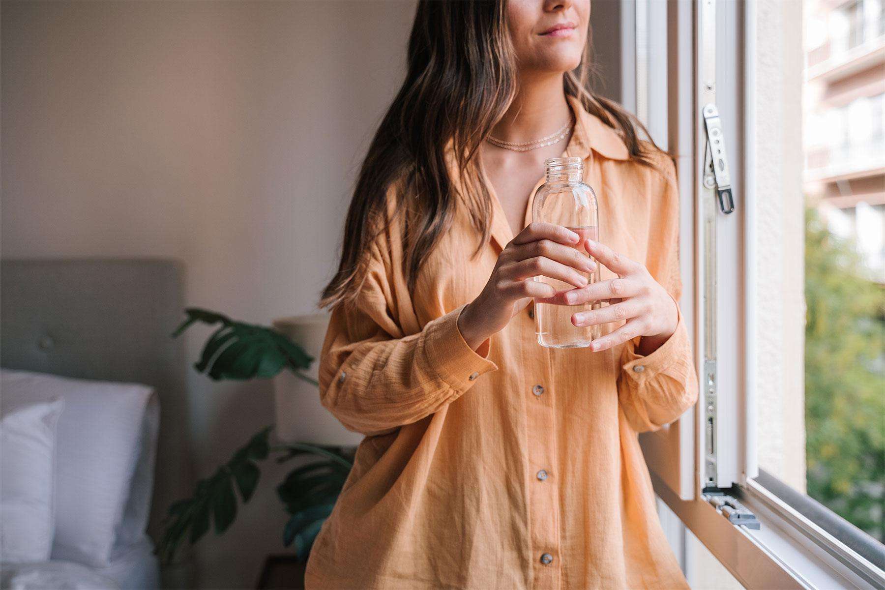 Woman leaning against window looking outside while holding a glass of water.