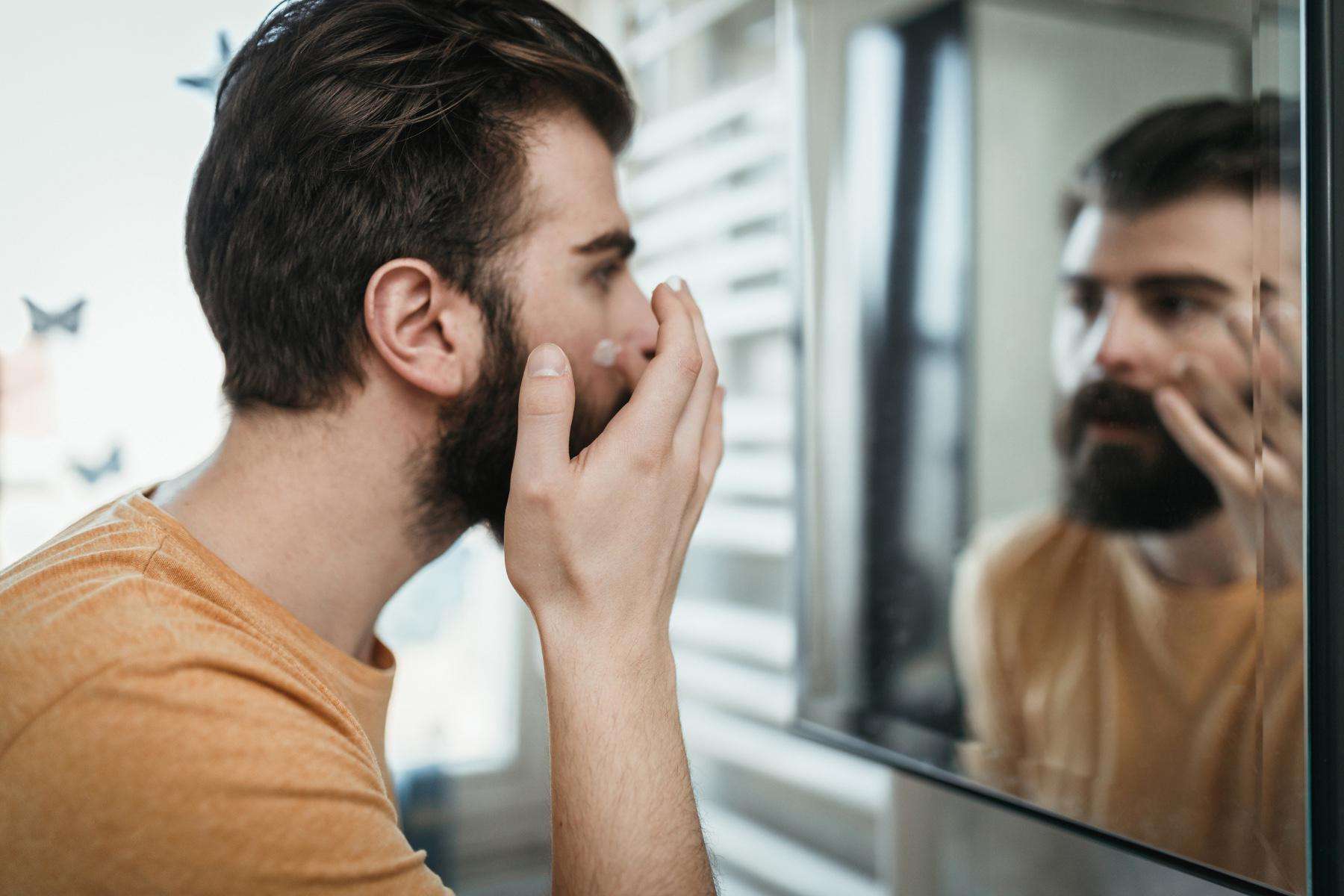 A man putting lotion on his face while looking into his bathroom mirror