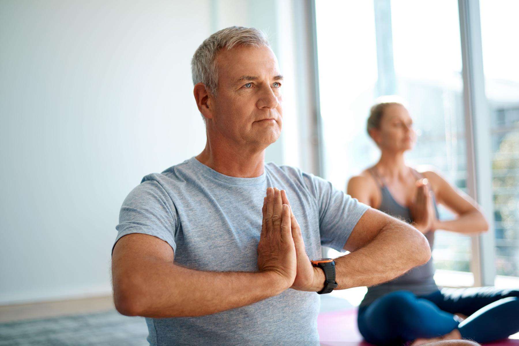 Close-up of an older man with hands at heart center, older woman blurry in the background with hands also at heart center.