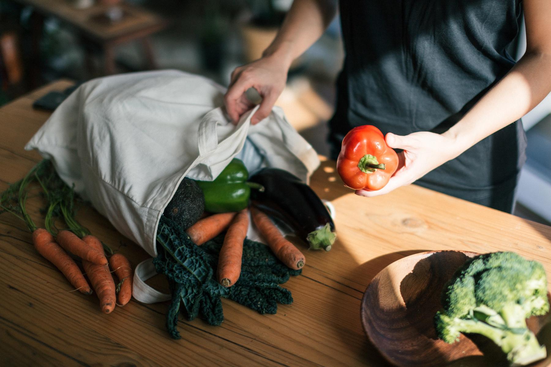 Close-up of hands putting a red pepper into a reusable canvas bag filled with fresh vegetables.