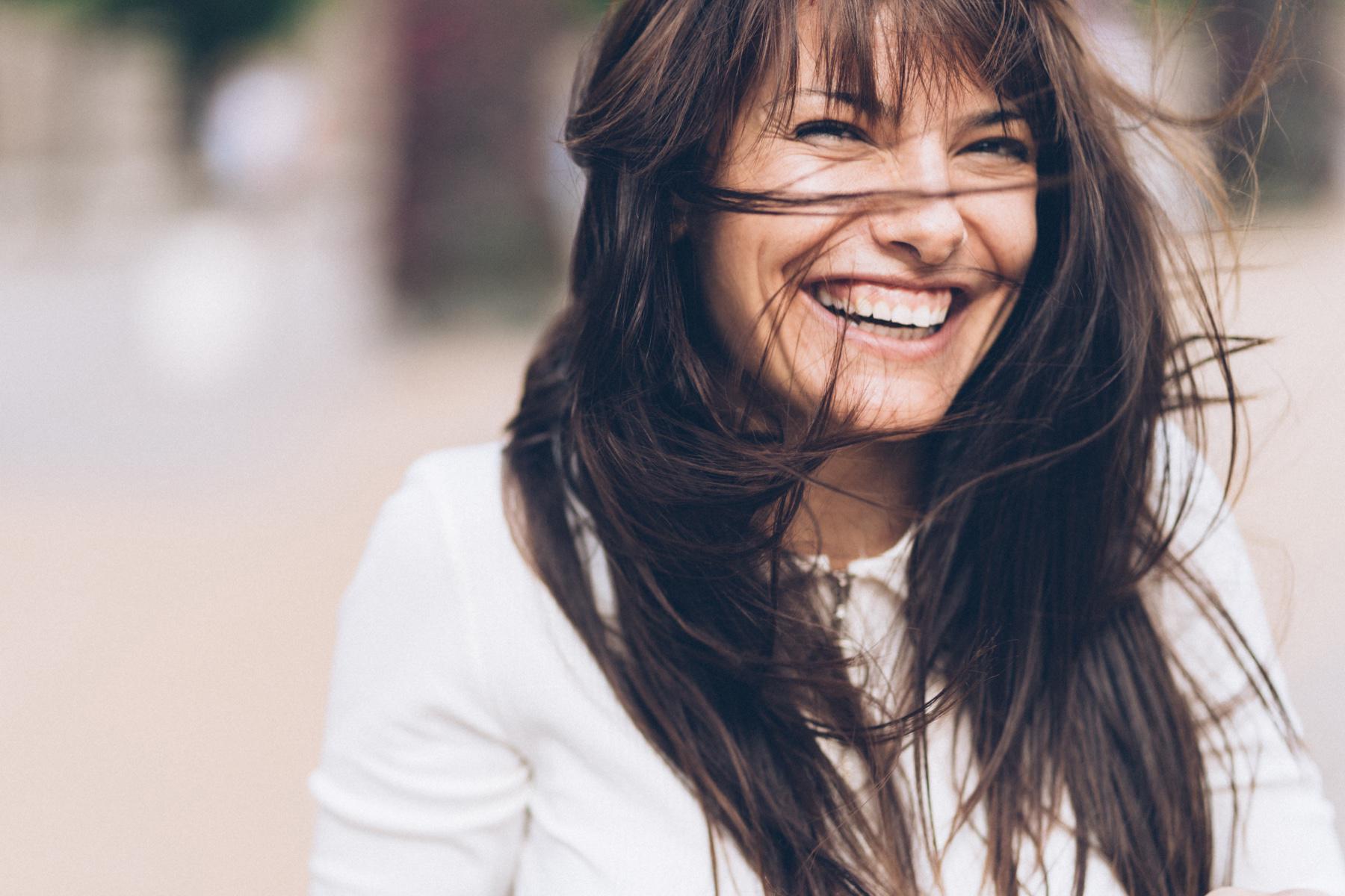 A brunette-haired woman laughing as her hair blows in the wind.