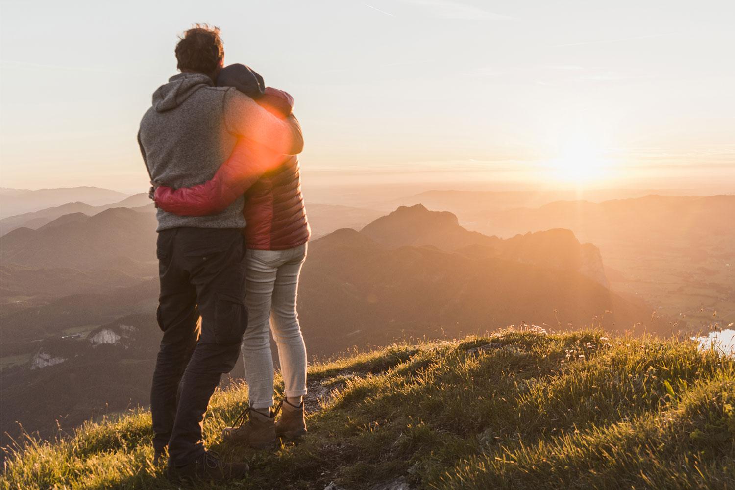 Man and woman standing on hillside together overlooking sunset.