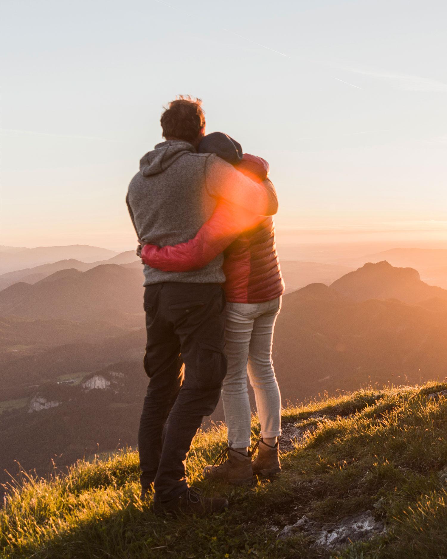 Man and woman standing on hillside together overlooking sunset.
